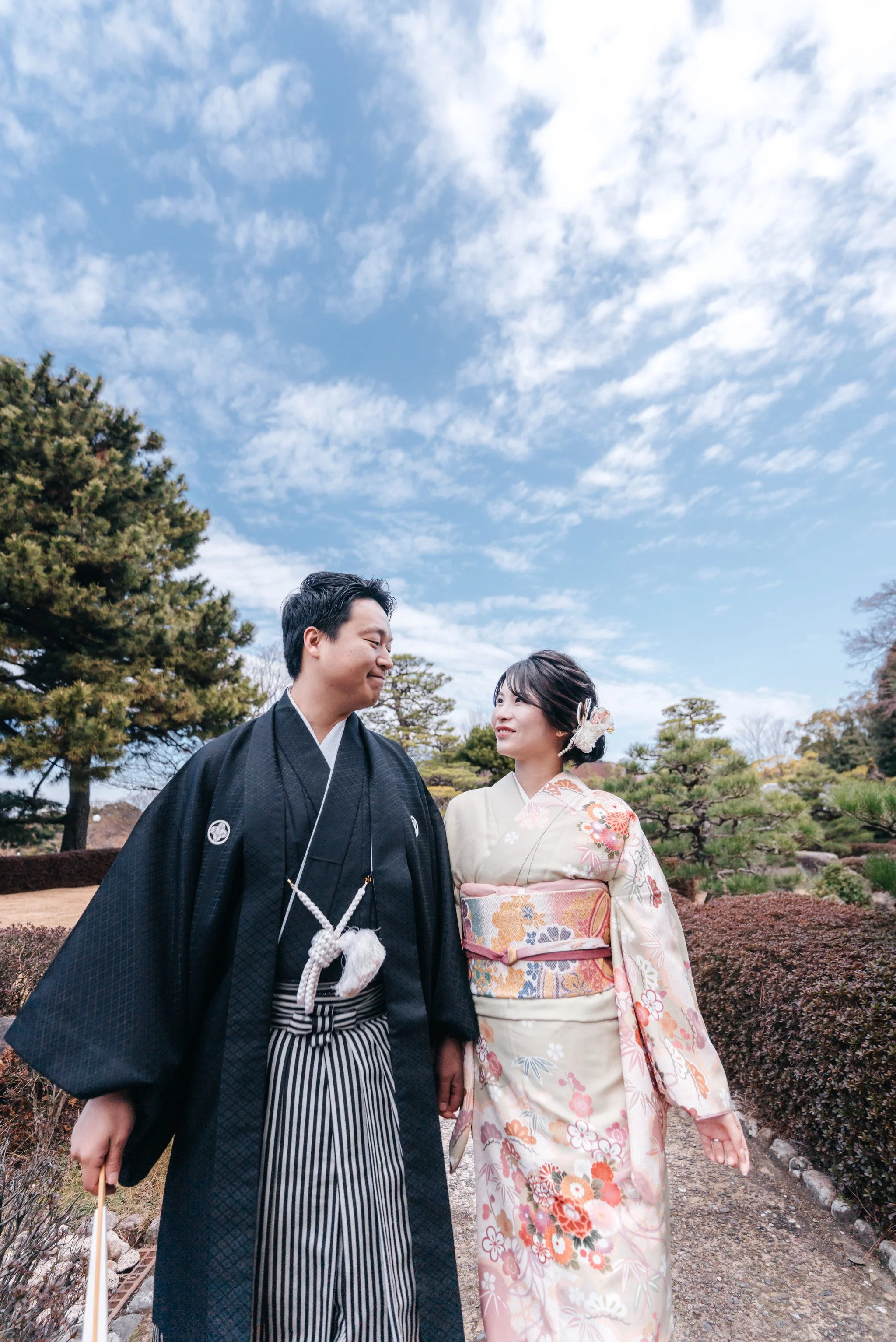 和装を着た若い夫婦が屋外で手をつなぎ、笑顔で見つめ合っています。青空と庭の木々が背景です。