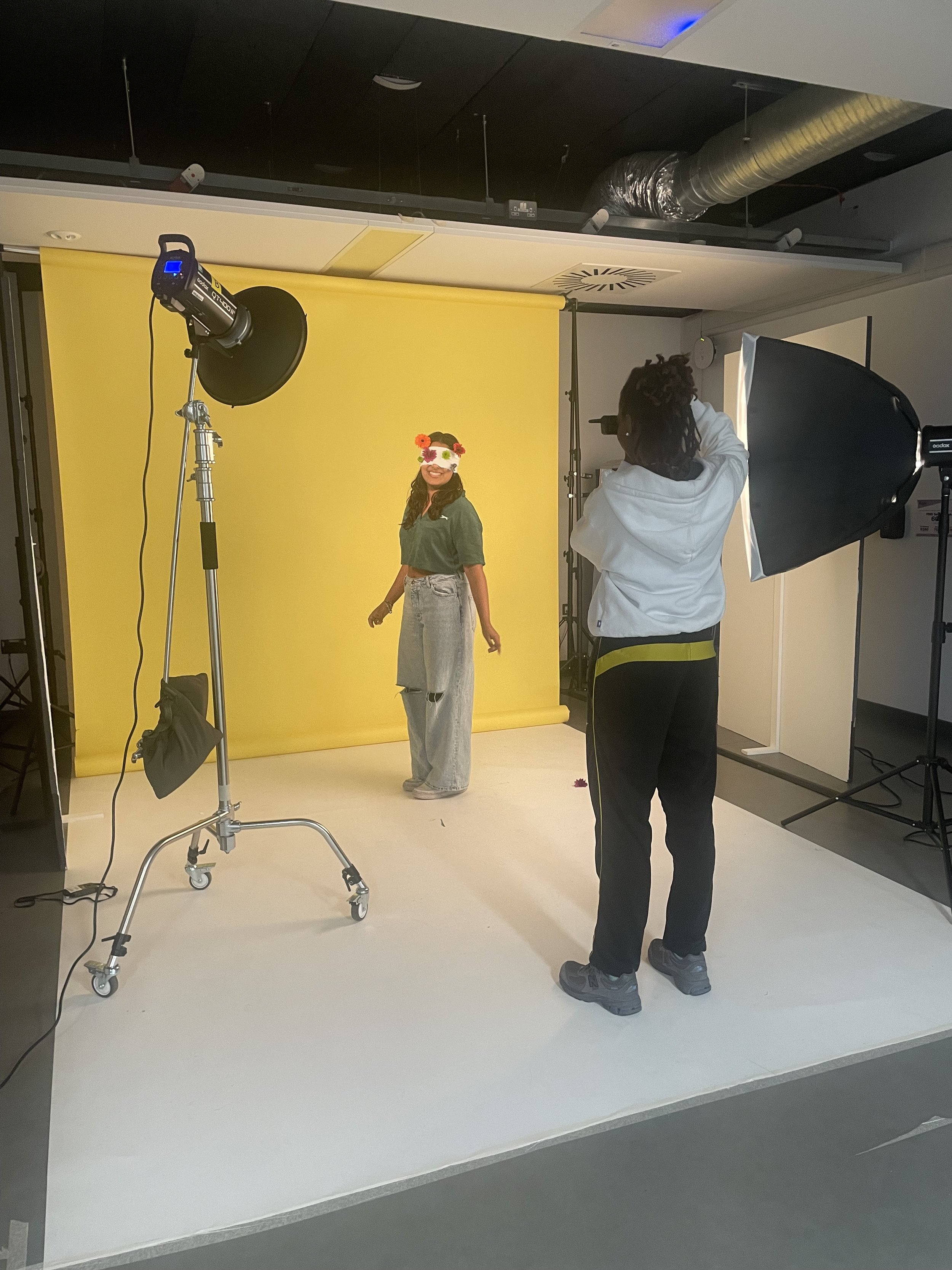 A woman is standing in front of a yellow backdrop in a photography studio, wearing a mask and a flower crown. A photographer is taking her picture with studio lights set up around her.