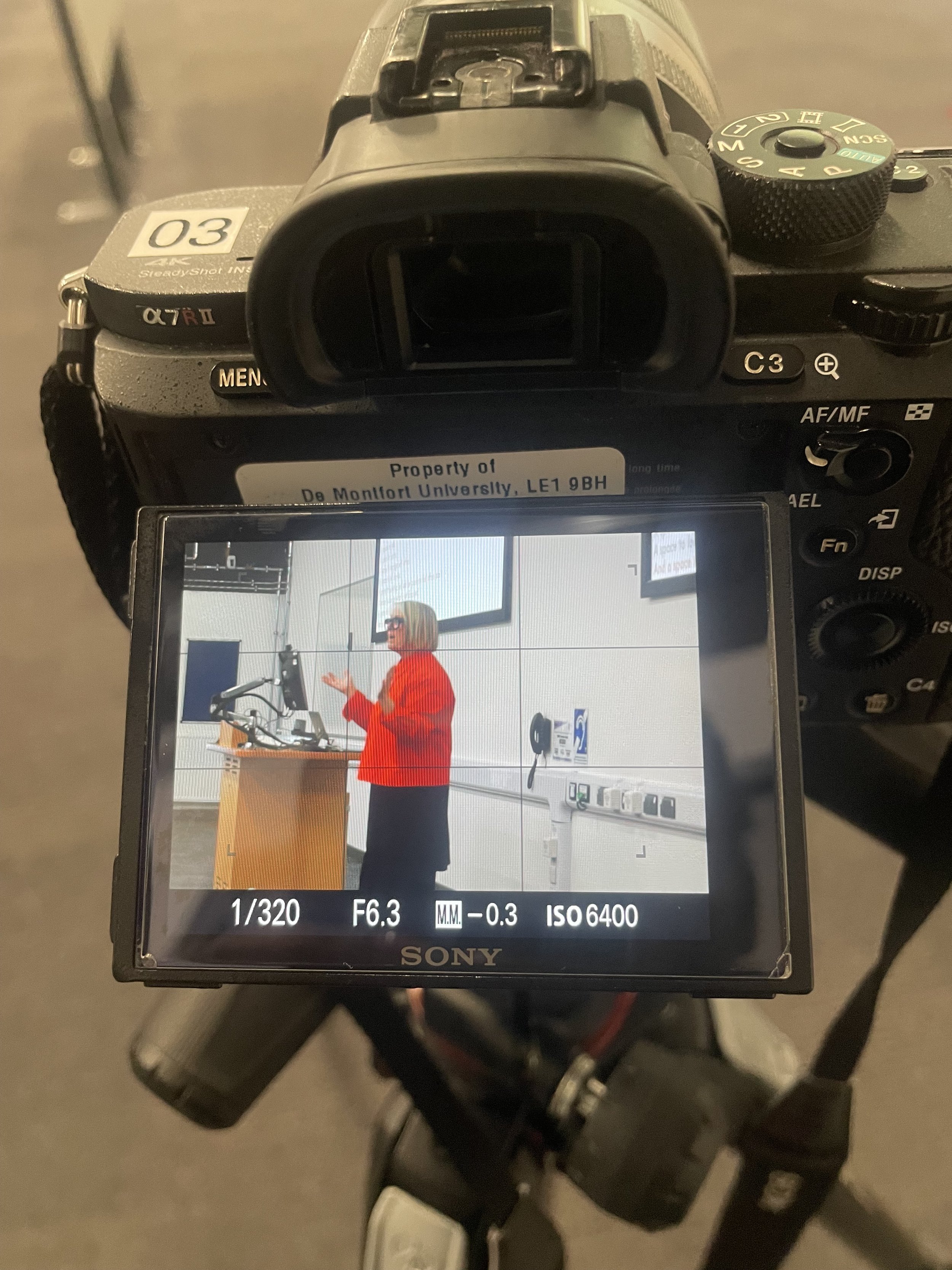 Photo of a digital camera capturing a woman in red at a podium, with the camera screen showing her speaking and gesturing, at a presentation or lecture event.