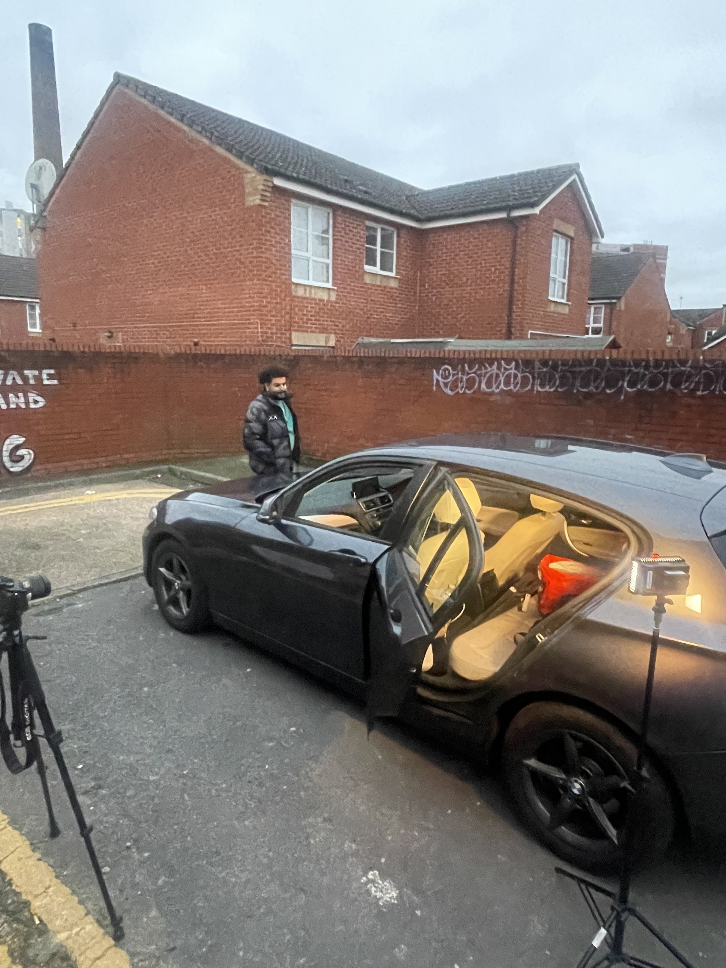 A black car with one open door parked on the street, a man standing nearby, and cameras set up for filming against a red brick wall and residential buildings.