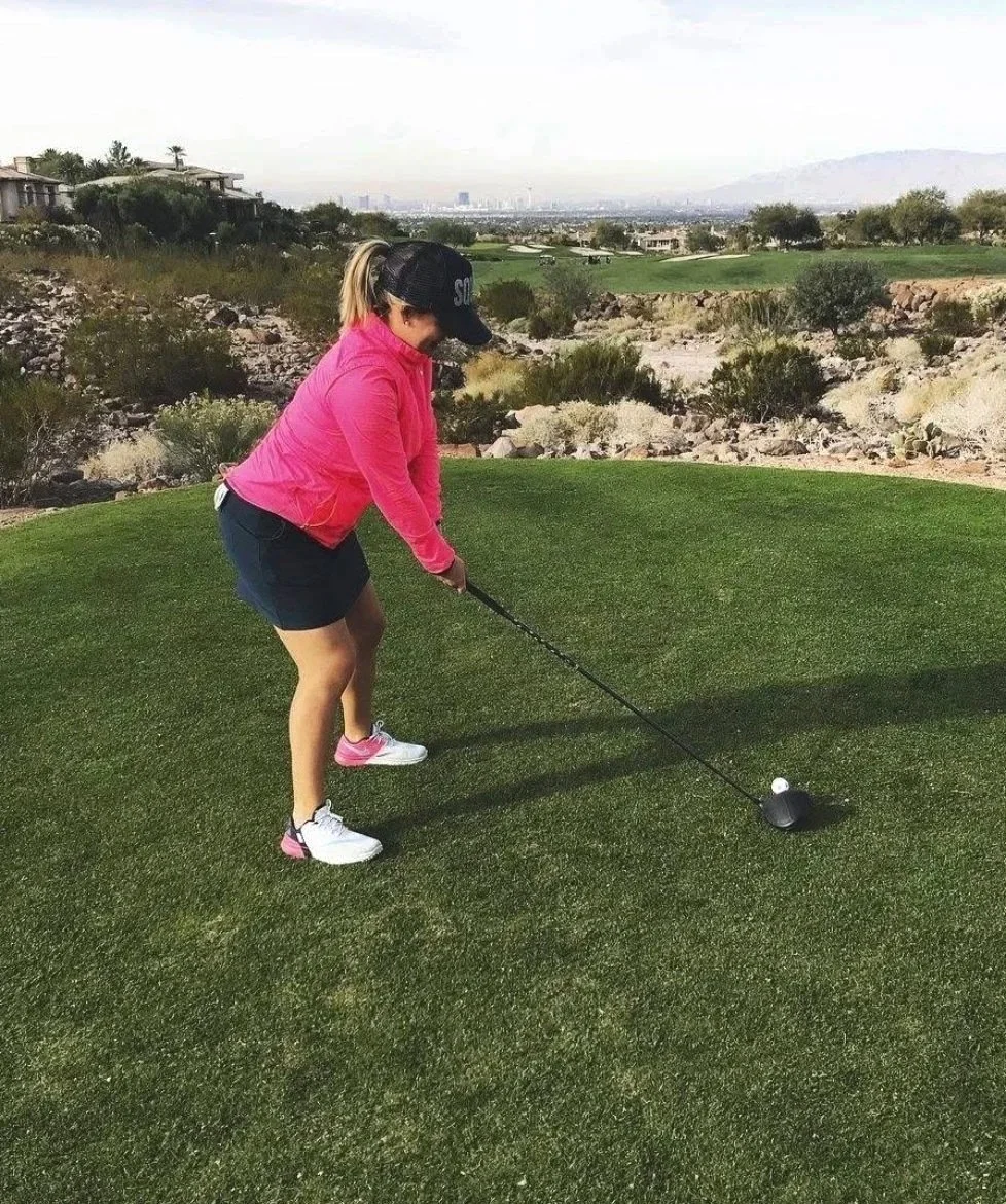 A woman in pink jacket and black cap prepares to tee off on a golf course, with cityscape and desert terrain in the background.