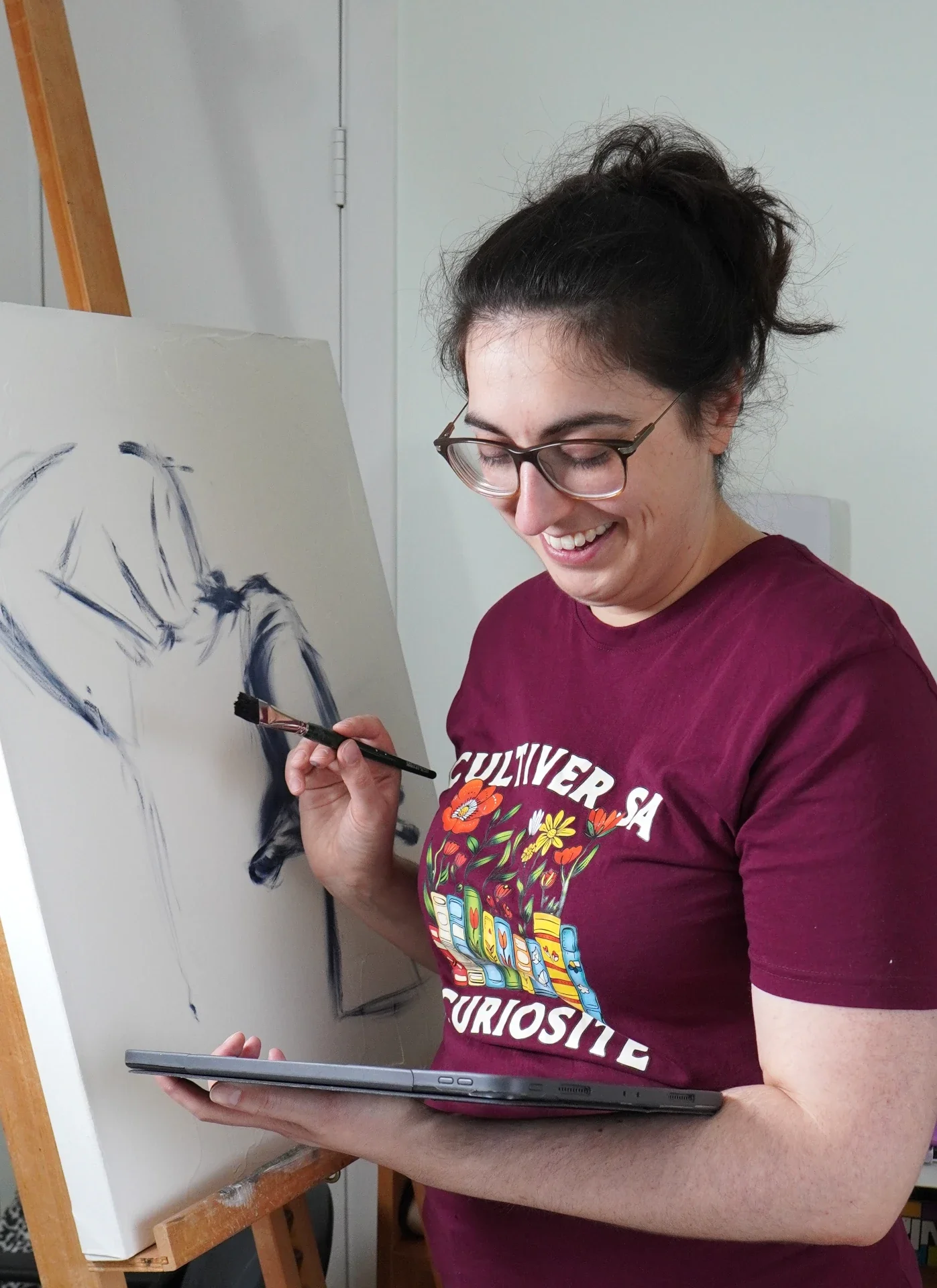 Woman painting a portrait on a canvas with a tablet in her hand, smiling and wearing a maroon t-shirt with colorful text and illustrations.