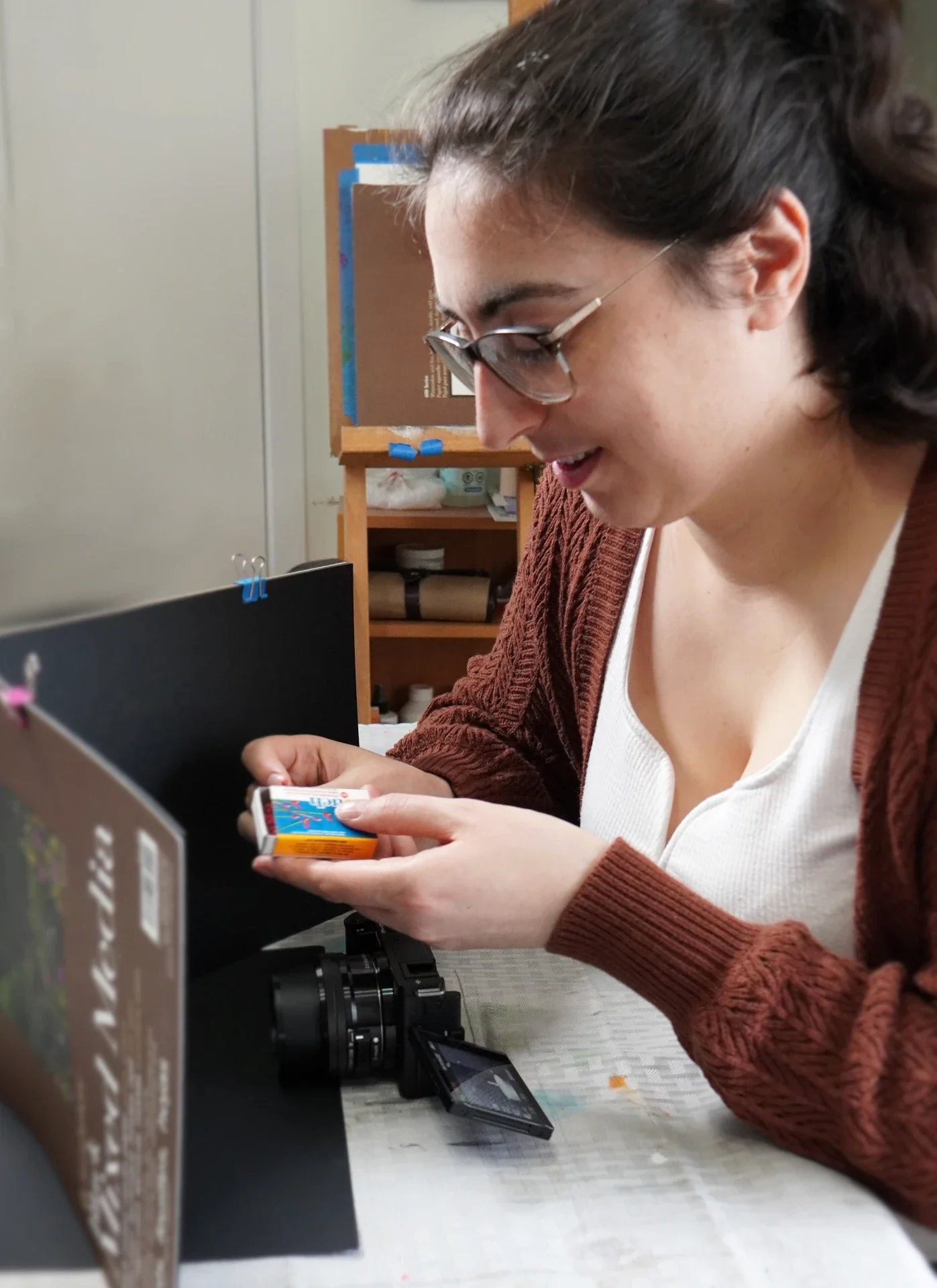 A woman with glasses and dark hair in a ponytail smiling and looking down at a pack of gum she is holding. She is sitting at a desk with a camera and a black computer monitor, against a background of a wooden shelf with books and objects.