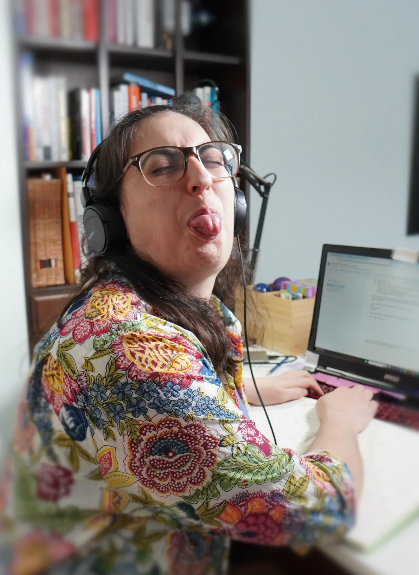 A woman with glasses and headphones making a silly face with her tongue out, sitting at a desk with a laptop.