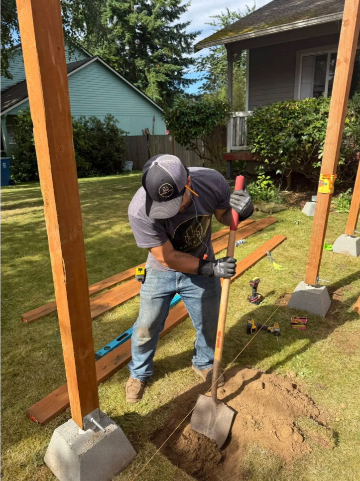 A man digging a hole in a backyard with a shovel during a construction or woodworking project, with wooden beams and construction tools surrounding him.