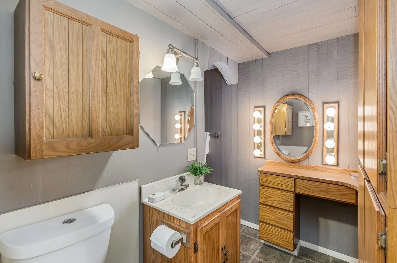 Bathroom with wooden cabinets, a marble countertop with a small plant, a mirror, and light fixtures with round bulbs. The wall behind the vanity is gray paneling, and there is a toilet and sink.