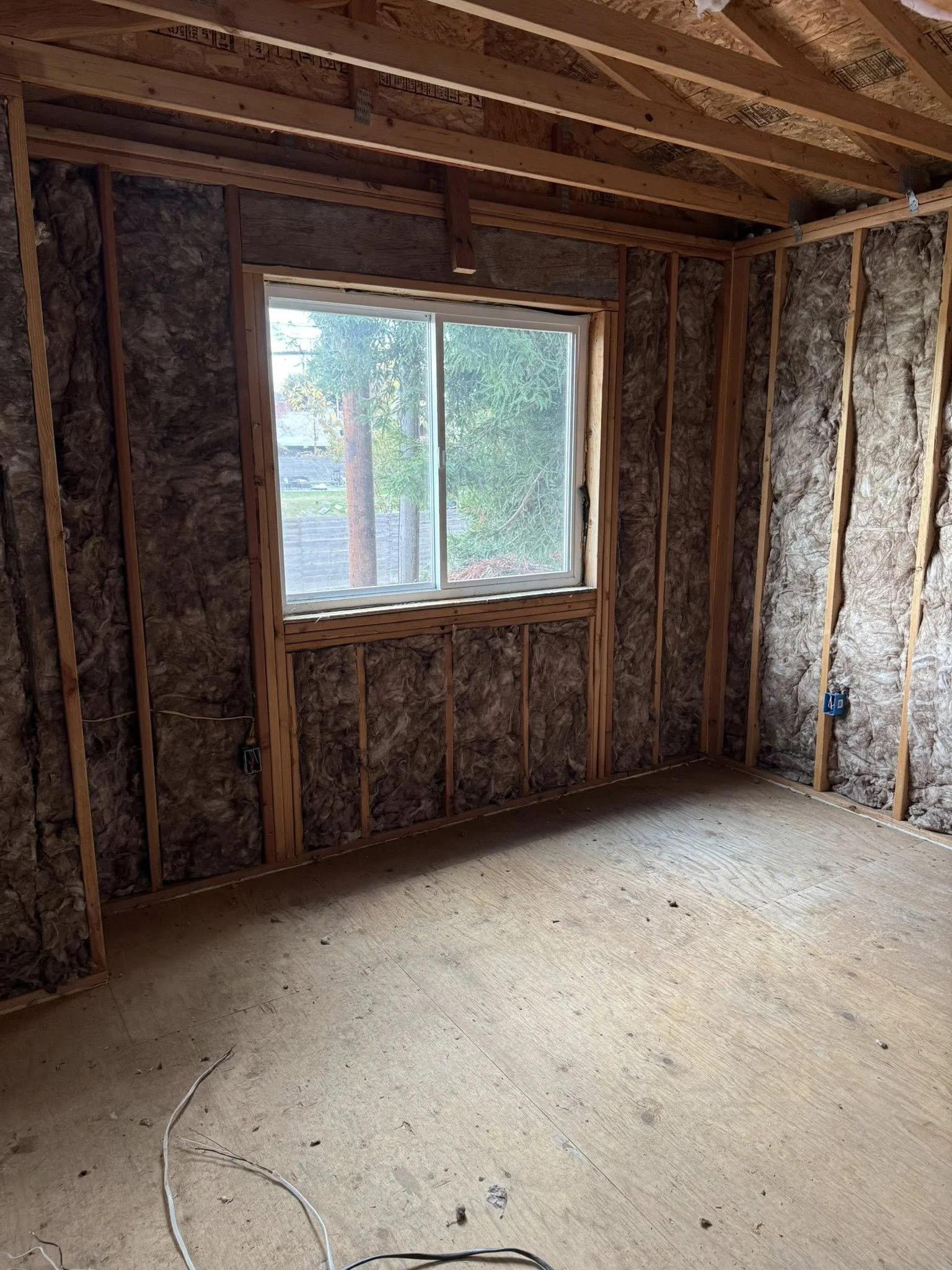 Interior of a room under construction with exposed insulation, wooden framing, and a window revealing trees outside.