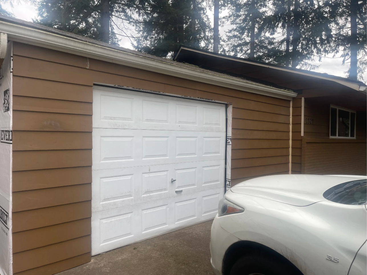 Front view of a house with brown siding, a white garage door, a window, and a white car parked in front. There is construction or renovation work on the garage door area.
