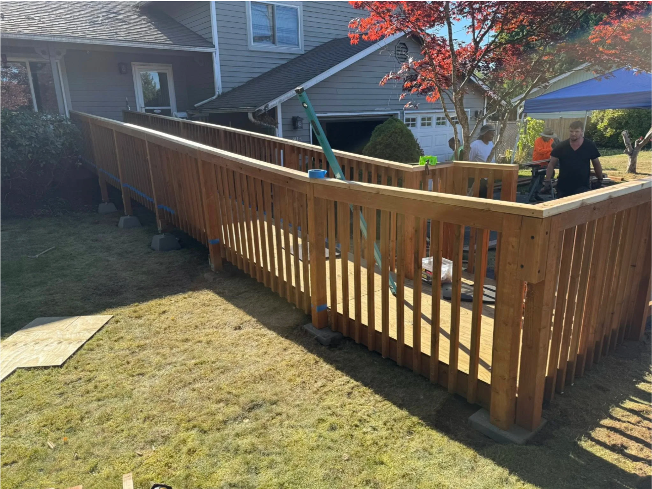 A newly built wooden deck with a railing being constructed in a backyard, with people working under a blue canopy and a tree with red leaves nearby.