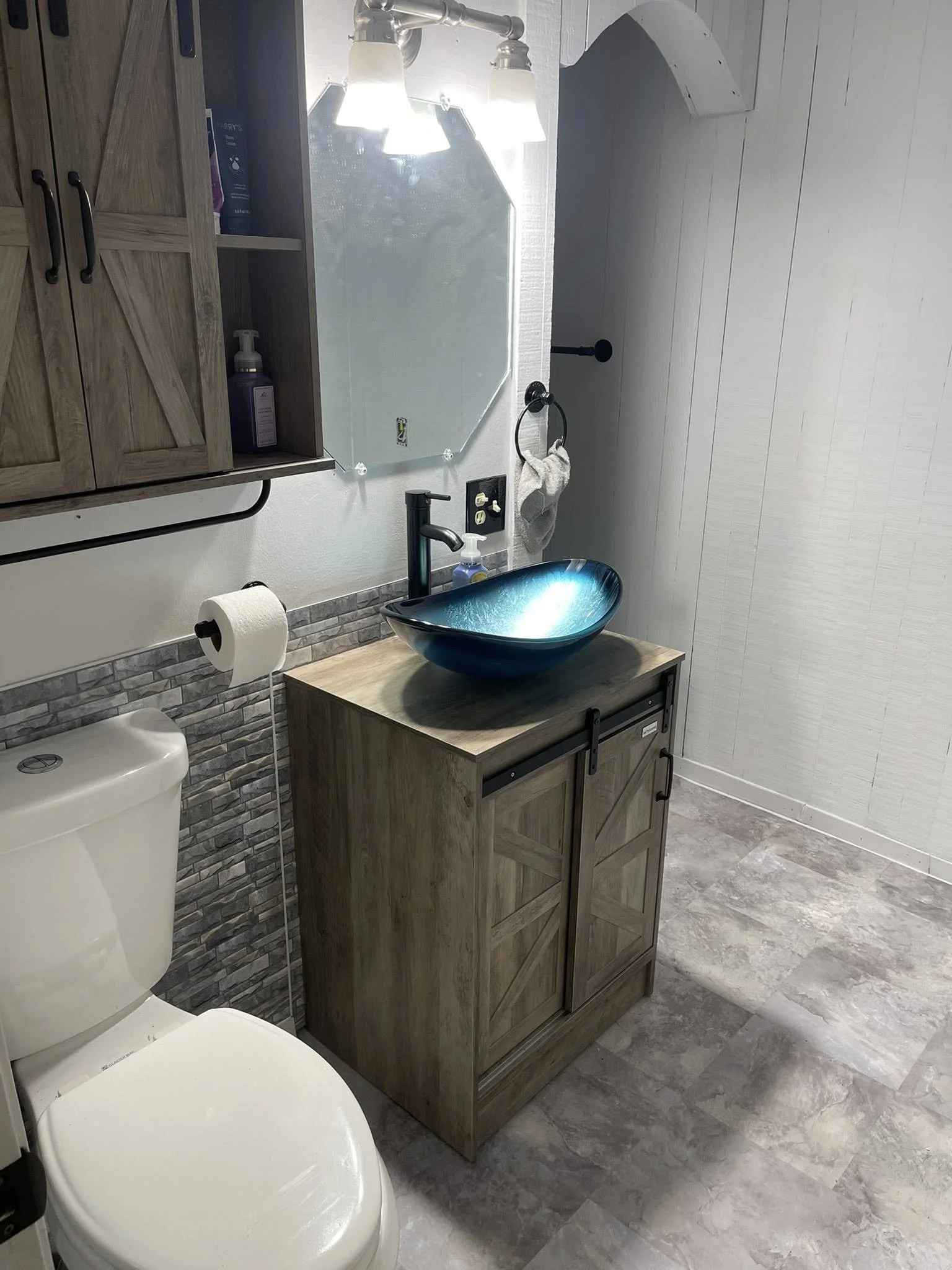 A bathroom with a toilet, a small wooden vanity with a blue glass vessel sink, a mirror, and a wall-mounted cabinet. The sink has a black faucet, and there is a paper towel roll next to the toilet. The walls are white and decorated with a gray stone backsplash behind the toilet.