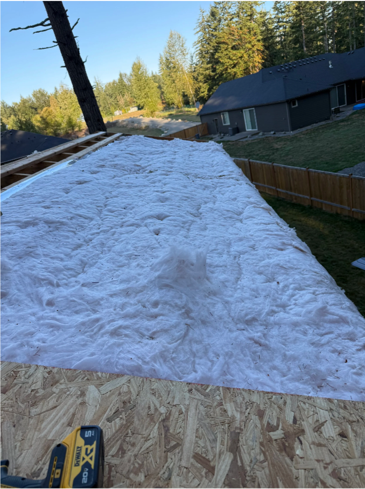 A construction site on a roof with white insulation material, wooden framing, and a cordless drill in the foreground. Houses and trees are visible in the background.