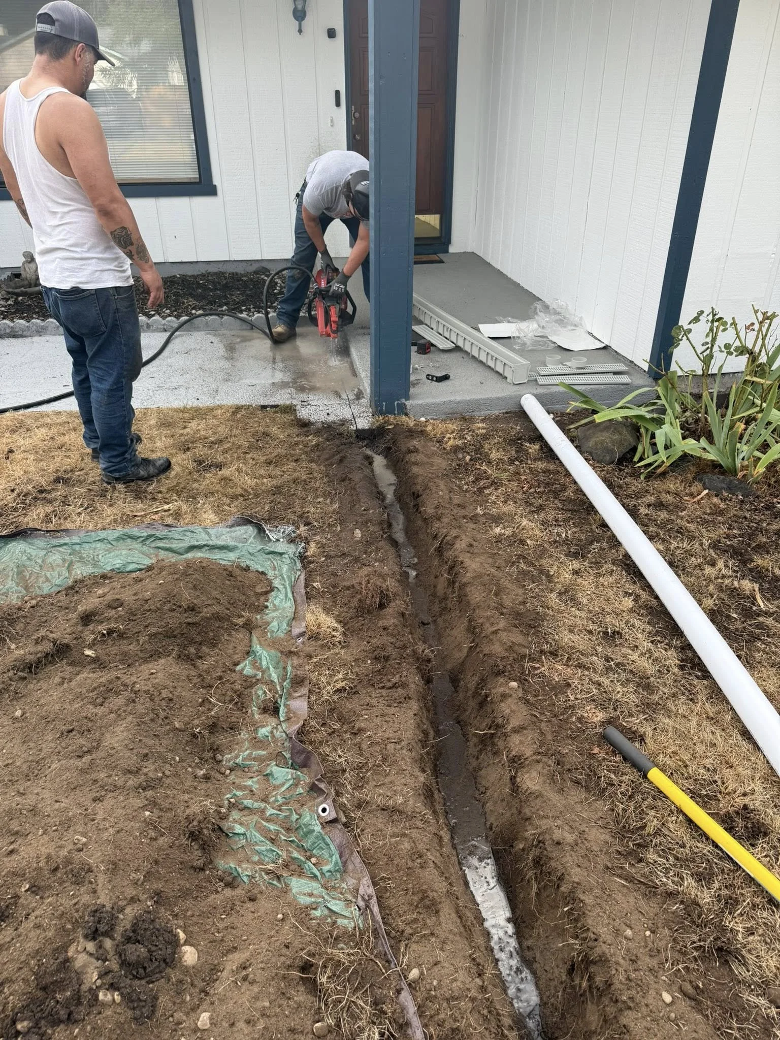 Two workers installing or repairing a drainage or utility line outside a house, with one using a concrete saw and the other overseeing, near the front porch.