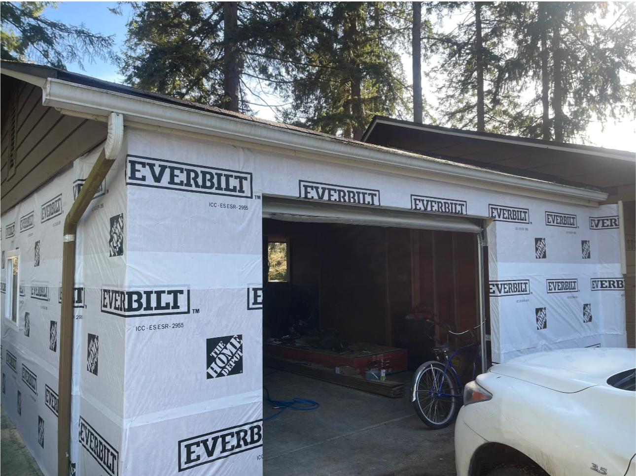 Garage under construction with white weather-resistant sheathing marked 'EVERBILT' and 'The Home Depot.' A bicycle and white car are parked outside. Some tools and materials are visible inside the garage.