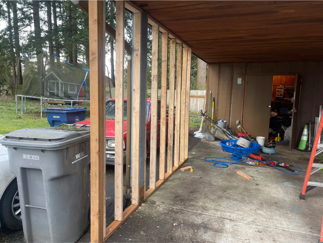 Garage under construction with wooden framing, tools, and supplies inside and outside, including garbage cans, a car, and a trampoline in the yard.