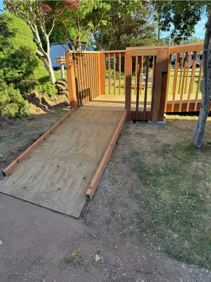 Wooden ramp leading up to a small deck with a railing, situated in a grassy backyard with trees.