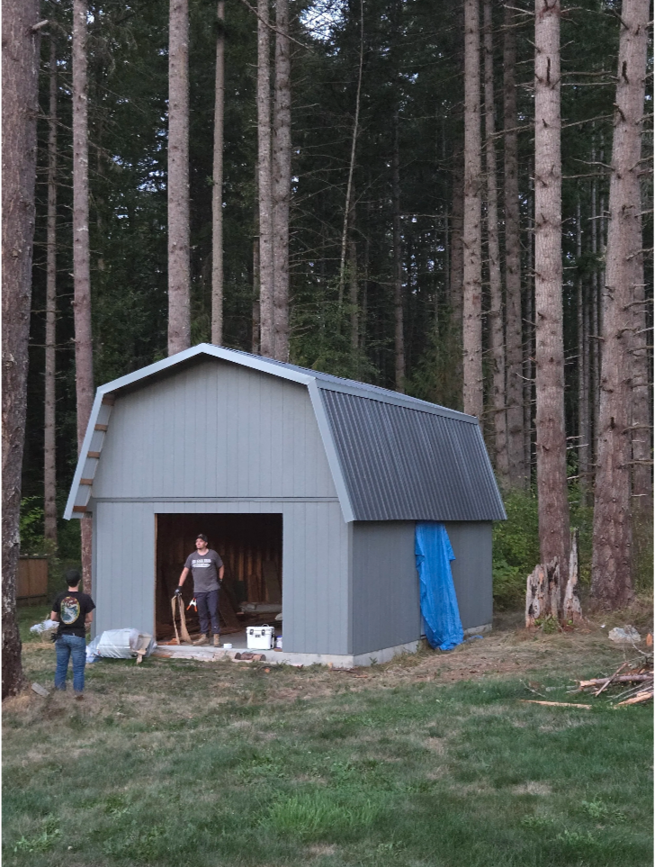 People working on a gray barn with a partially open door in a wooded area, with tall trees and some tools and materials around.