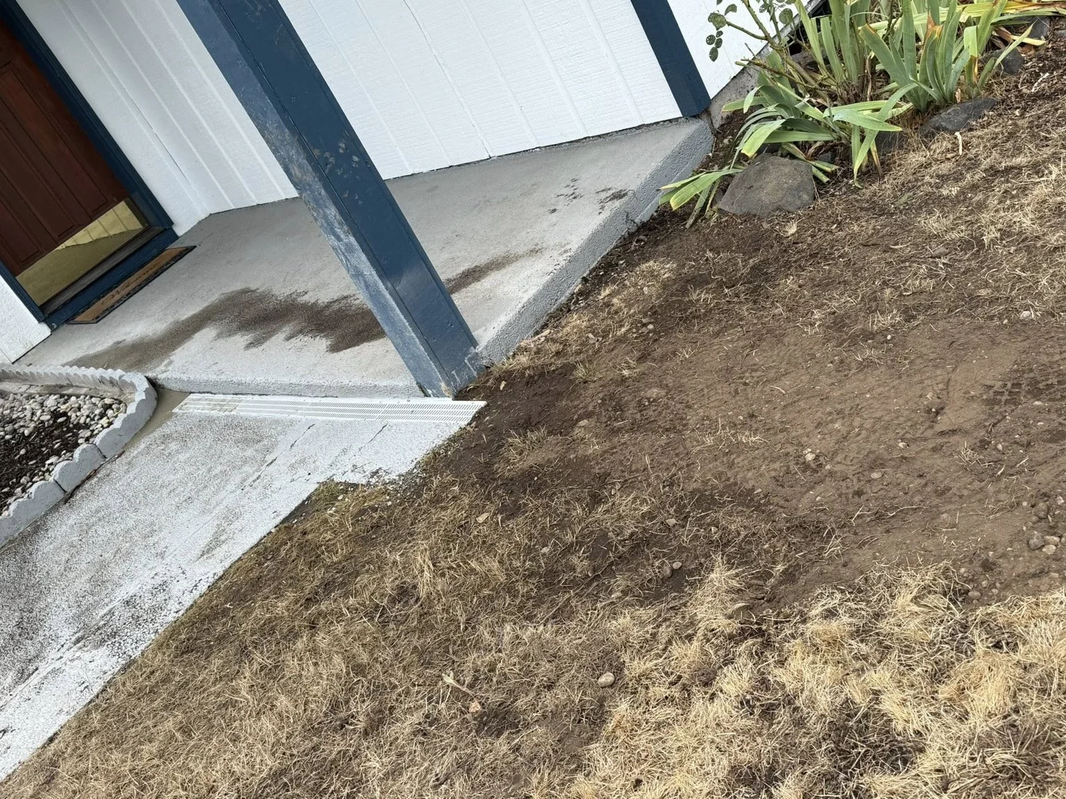 Part of a house exterior shows a concrete porch, a door with a welcome mat, and a small garden bed with green plants and rocks, with a patch of bare soil and dried grass in the yard.