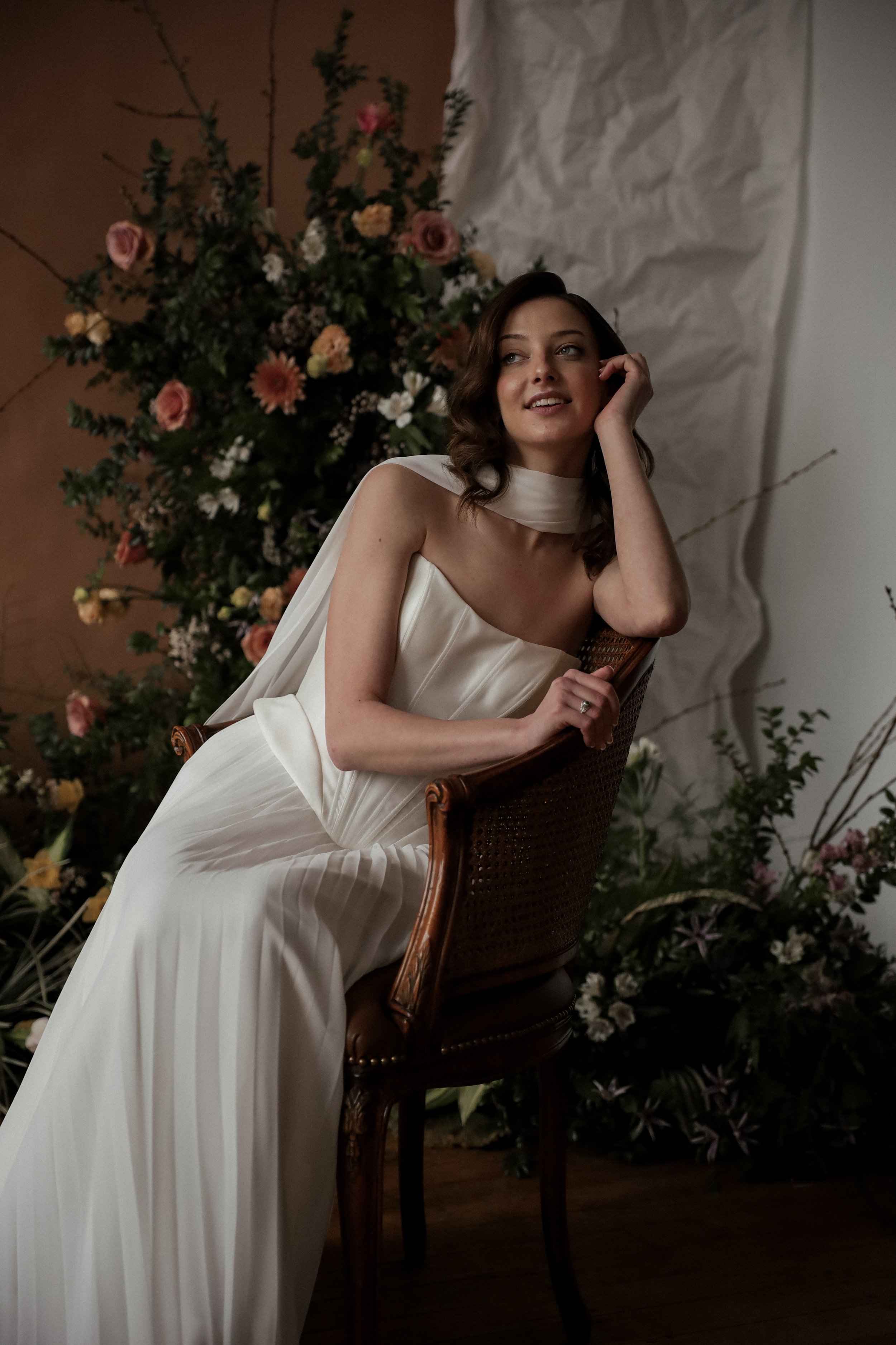 A woman in a white satin gown sitting on a wooden chair with floral arrangements in the background.