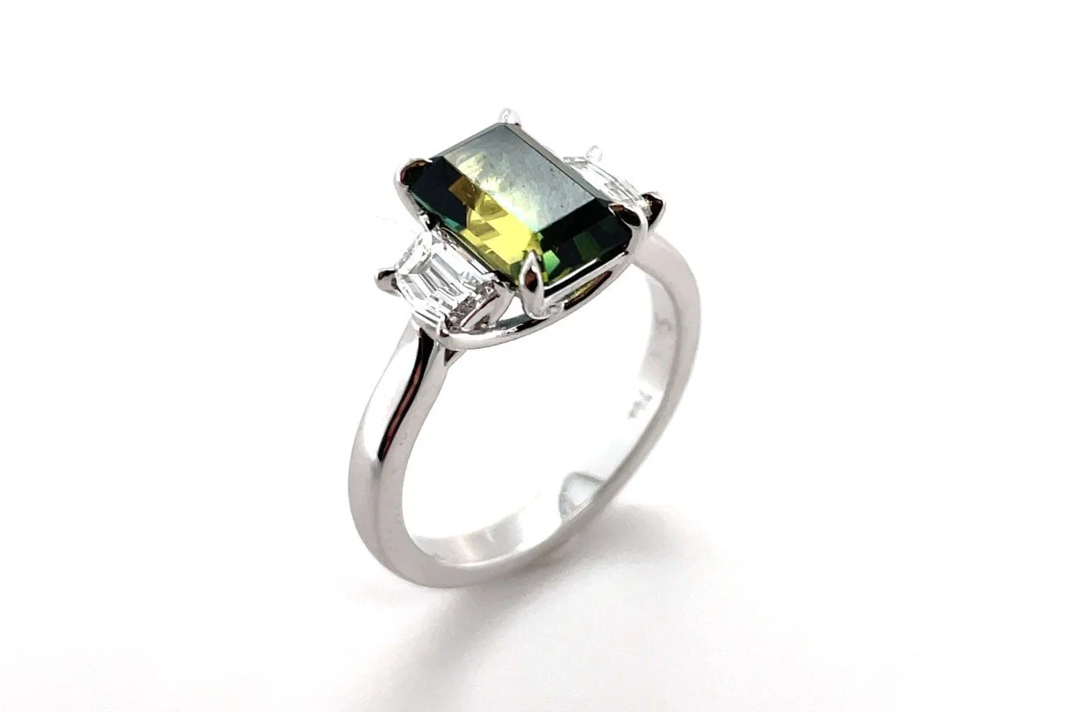 An angled top down view of a white gold ring with fancy cut diamonds either side of a green and yellow toned parti sapphire on white background