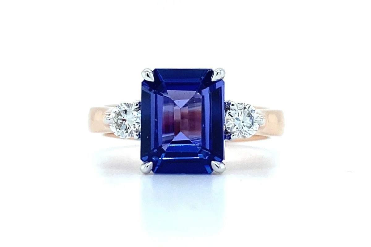 A front view of a trilogy ring with a purple emerald cut gemstone and two round white diamonds either side on a white background