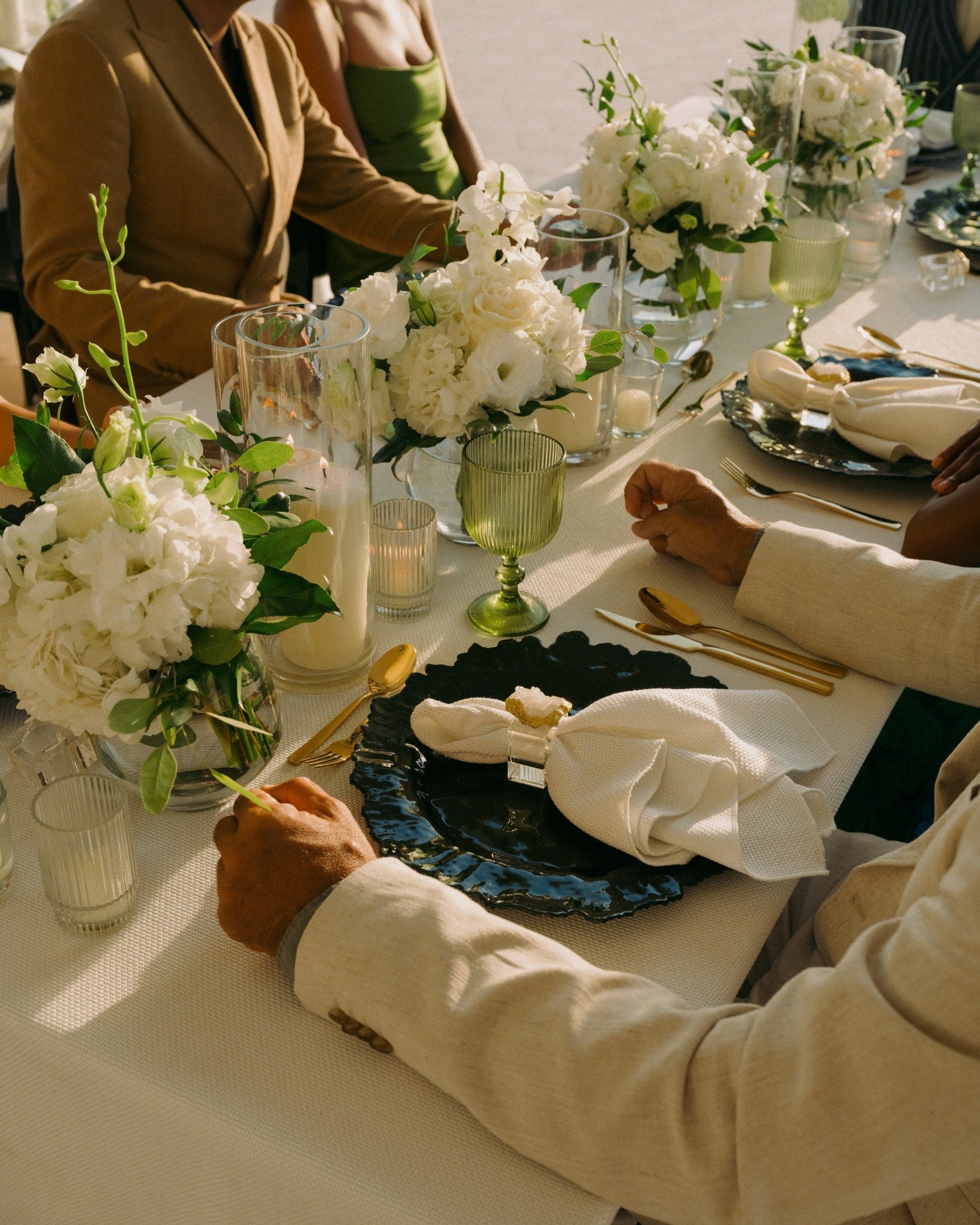 Elegant dinner table set with white floral arrangements, votive candles, green goblets, black and white plates, and gold utensils, with guests dressed in formal attire. Honeymoon, wedding, special travel.