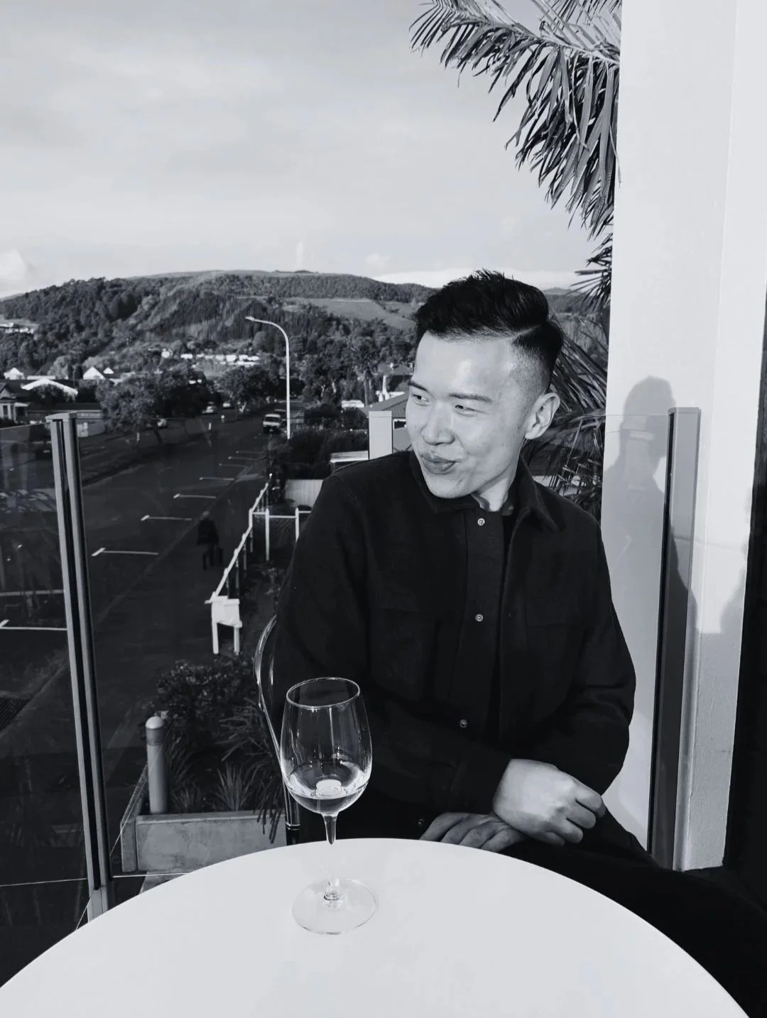 A young man with short dark hair, wearing a black jacket, sitting at a table with an empty wine glass in a restaurant or cafe, with a view of a parking lot, street, and hills. Ken Yao luxury travel advisor with Fora and Miharo Journeys.