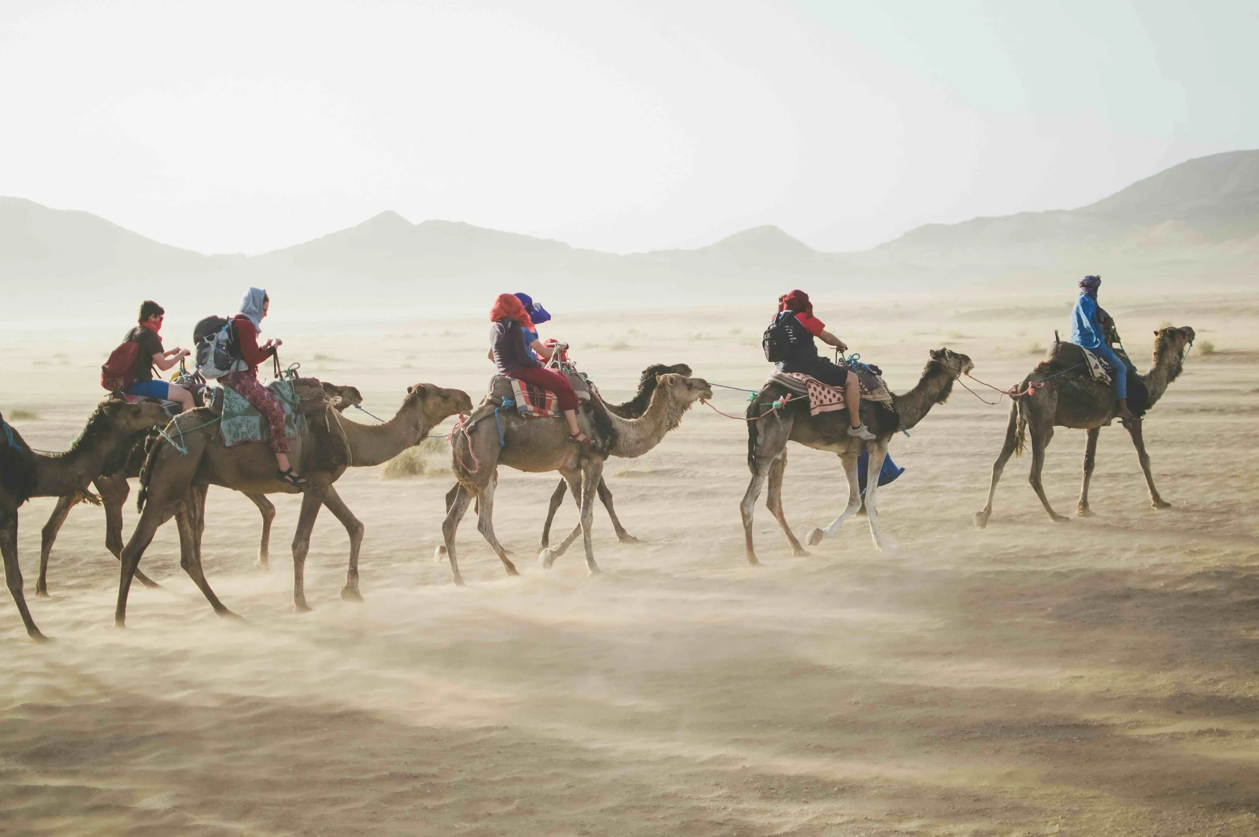 A group of five people riding camels in a desert landscape with mountains in the background.