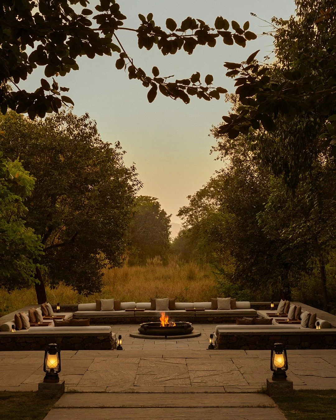 Outdoor seating area with a fire pit in the center, surrounded by sofas and cushions, at dusk in a natural wooded setting with trees and a grassy clearing. Aman resort luxury travel advisor, virtuoso.