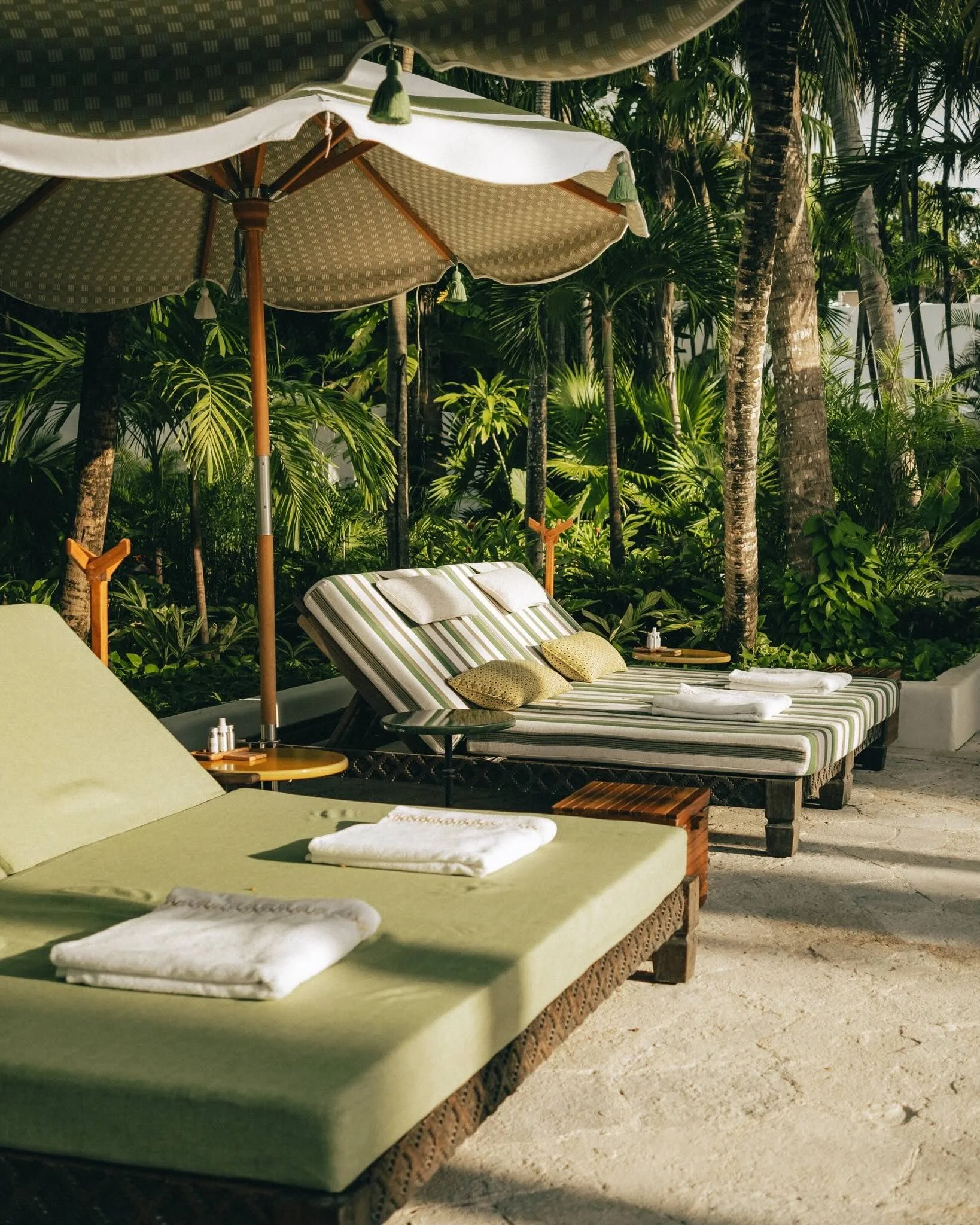 Two lounge chairs with cushions and towels, surrounded by tropical plants and trees, under large umbrellas on a sandy surface, in a tropical outdoor setting. Luxury travel advisor, virtuoso.