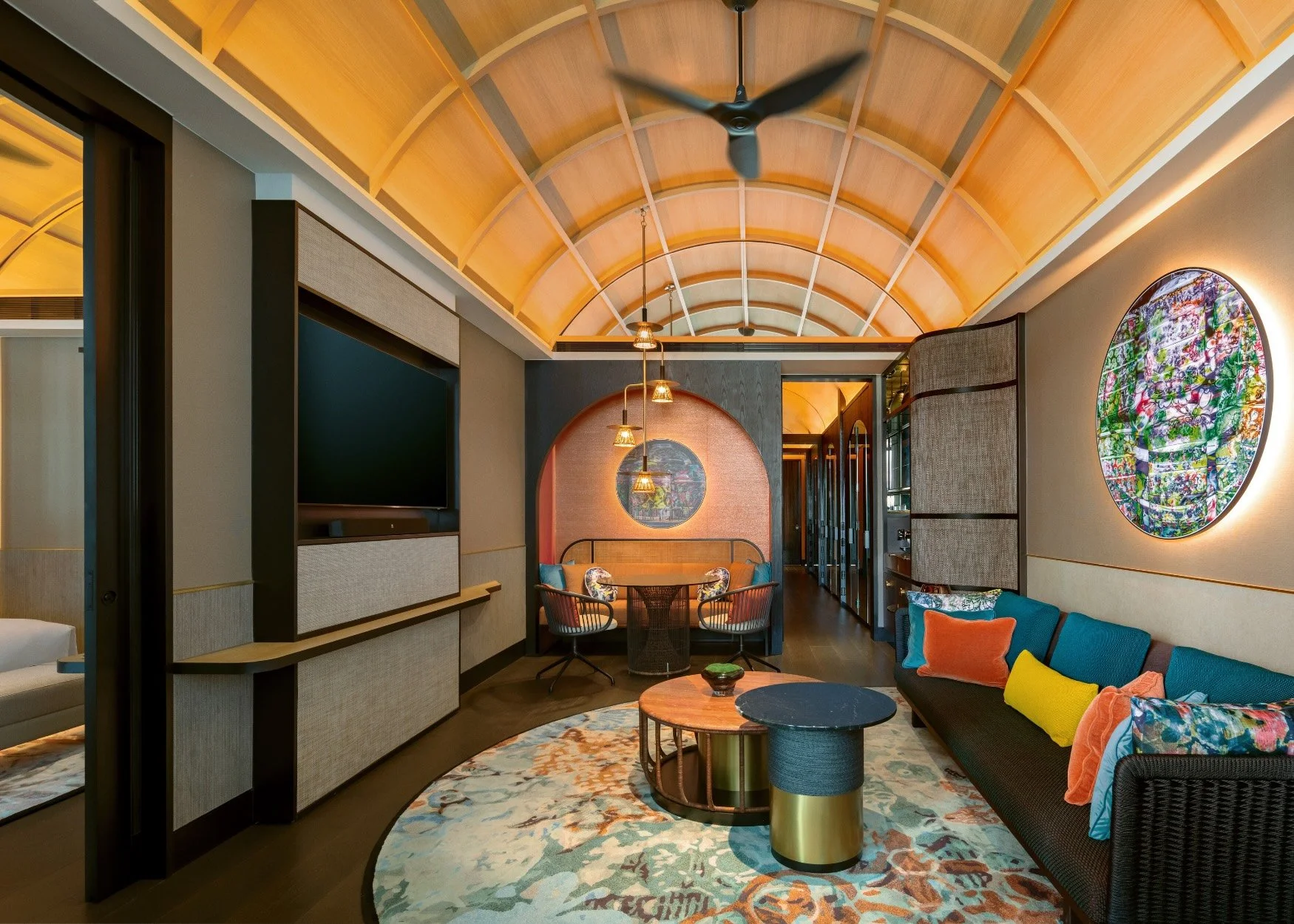 Interior of a modern living room with a curved wooden ceiling, a black ceiling fan, a flat-screen TV, colorful pillows, a circular rug, and a sitting area with chairs and a sofa.