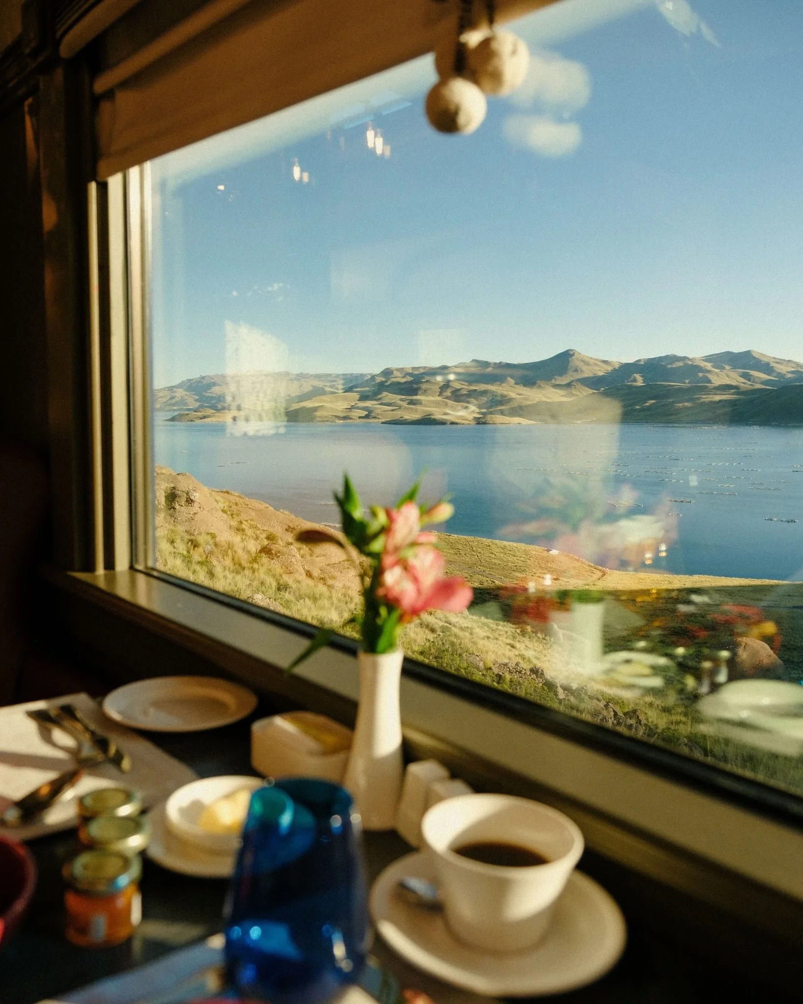 A window view of a lake and mountains with a breakfast table inside, including a vase with pink flowers, a cup of coffee, and small jars. Belmond oriental express luxury travel advisor, virtuoso.