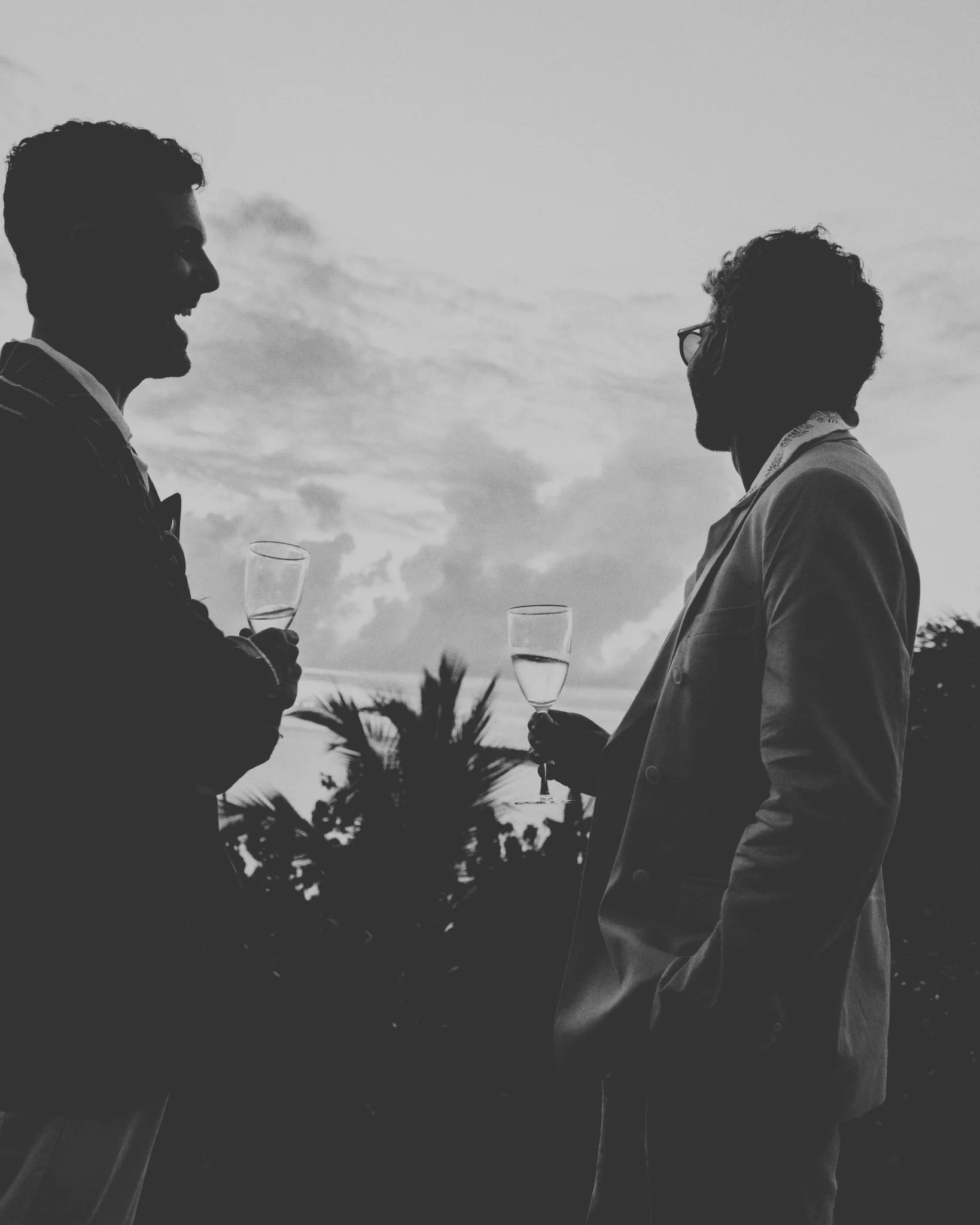 Silhouettes of two men in suits talking and holding glasses of champagne outdoors during sunset, with trees and cloudy sky in the background.