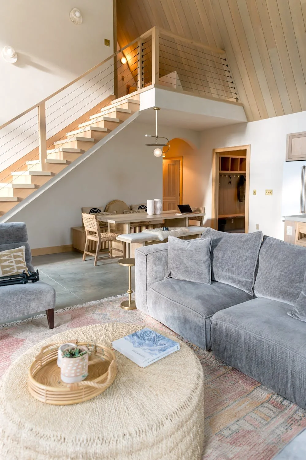 Living room with gray sofa, round woven ottoman with tray and book, dining table, and staircase with wooden steps and white railing