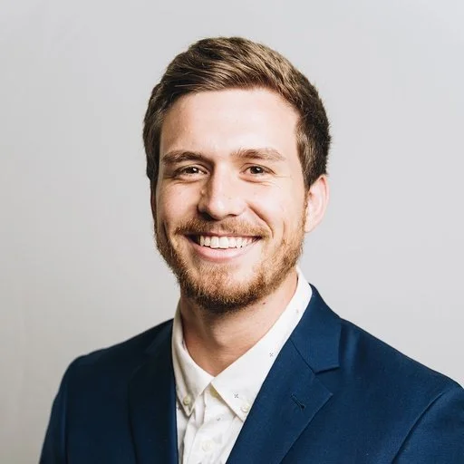 Portrait of a smiling man with light brown hair and beard, wearing a dark blue suit jacket and white shirt, against a plain light gray background.