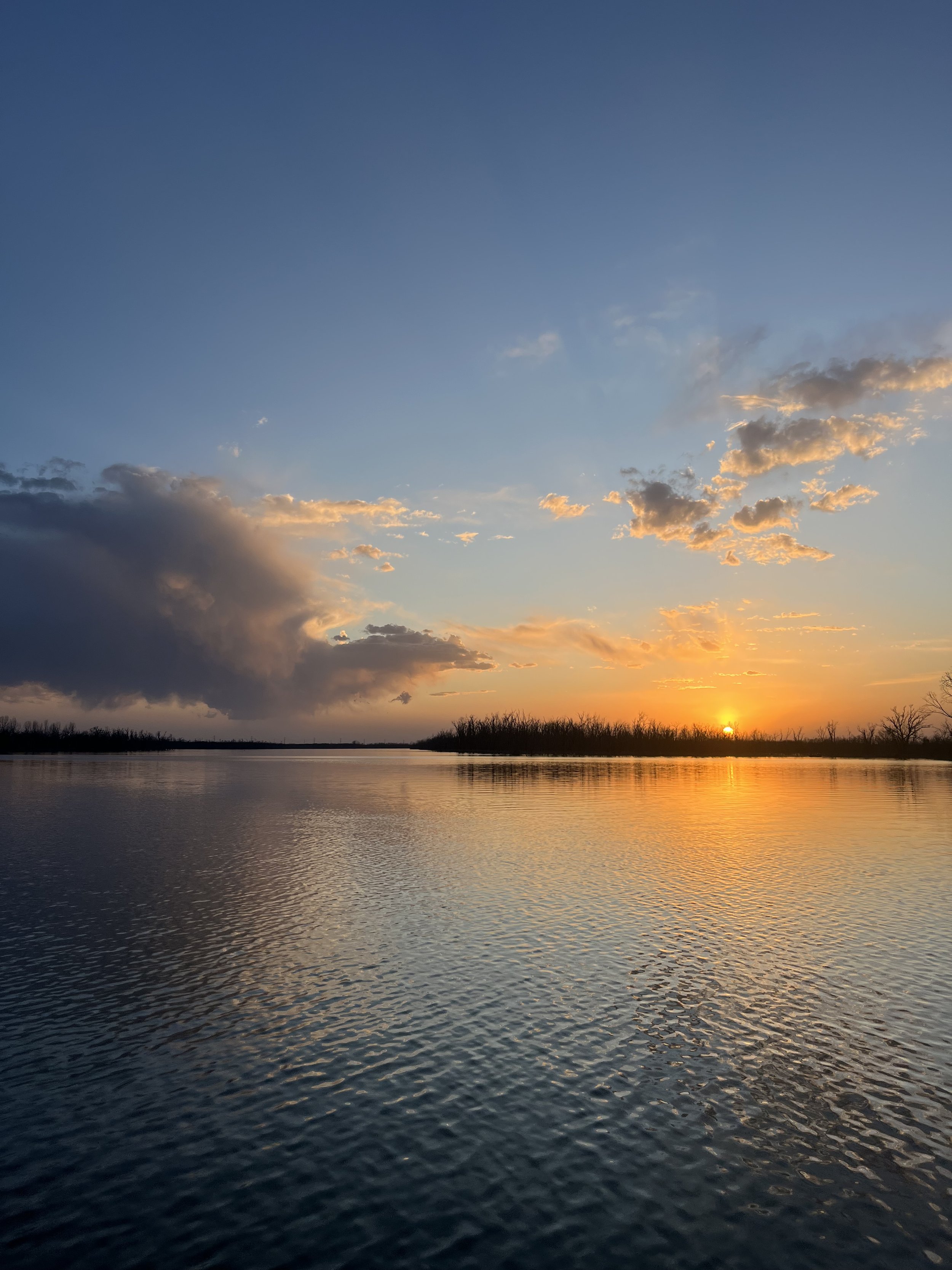Sunset over a calm river with a partly cloudy sky and silhouetted trees in the distance.