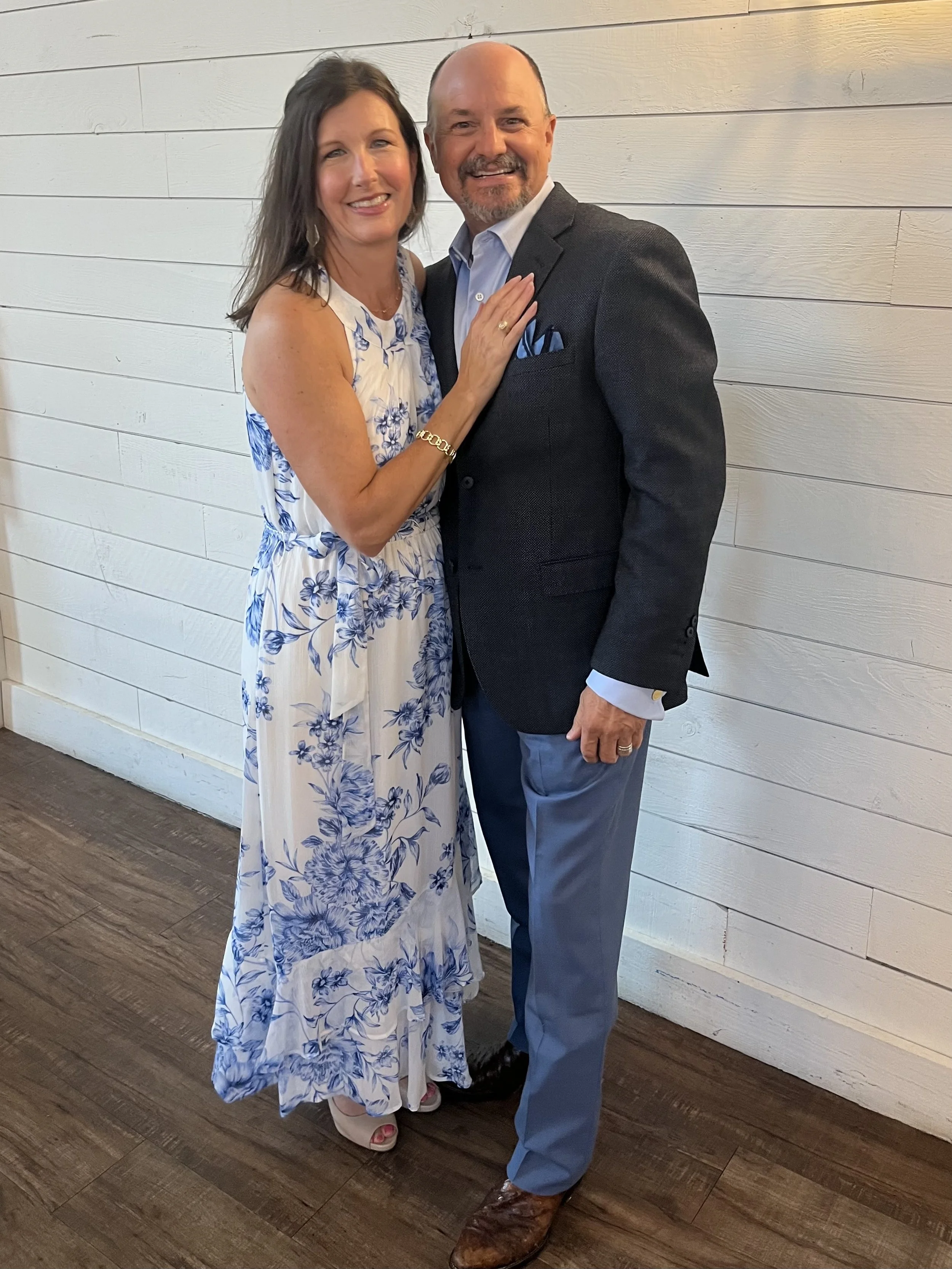 A smiling couple stands close together indoors against a white wooden wall. The woman wears a white and blue floral maxi dress and beige heels, while the man wears a dark blazer, light blue pants, and brown shoes.