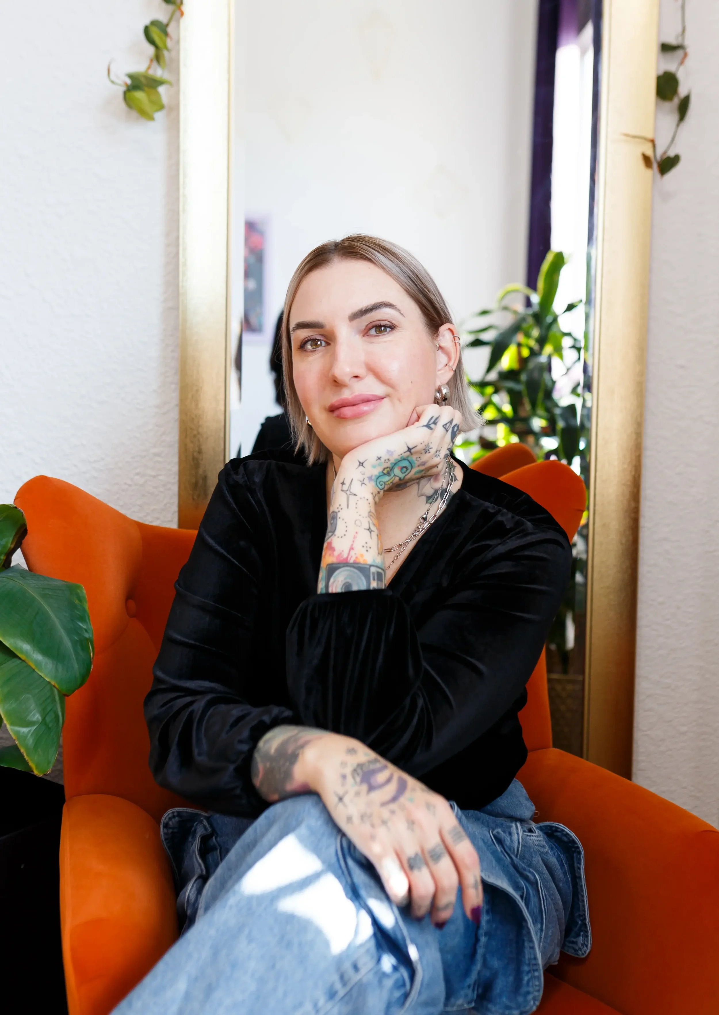 Artist Leah Crockford with tattoos, short brown hair, wearing a black velvet top and jeans, sitting on an orange chair in front of a mirror with a gold frame, smiling with her chin resting on her hand.