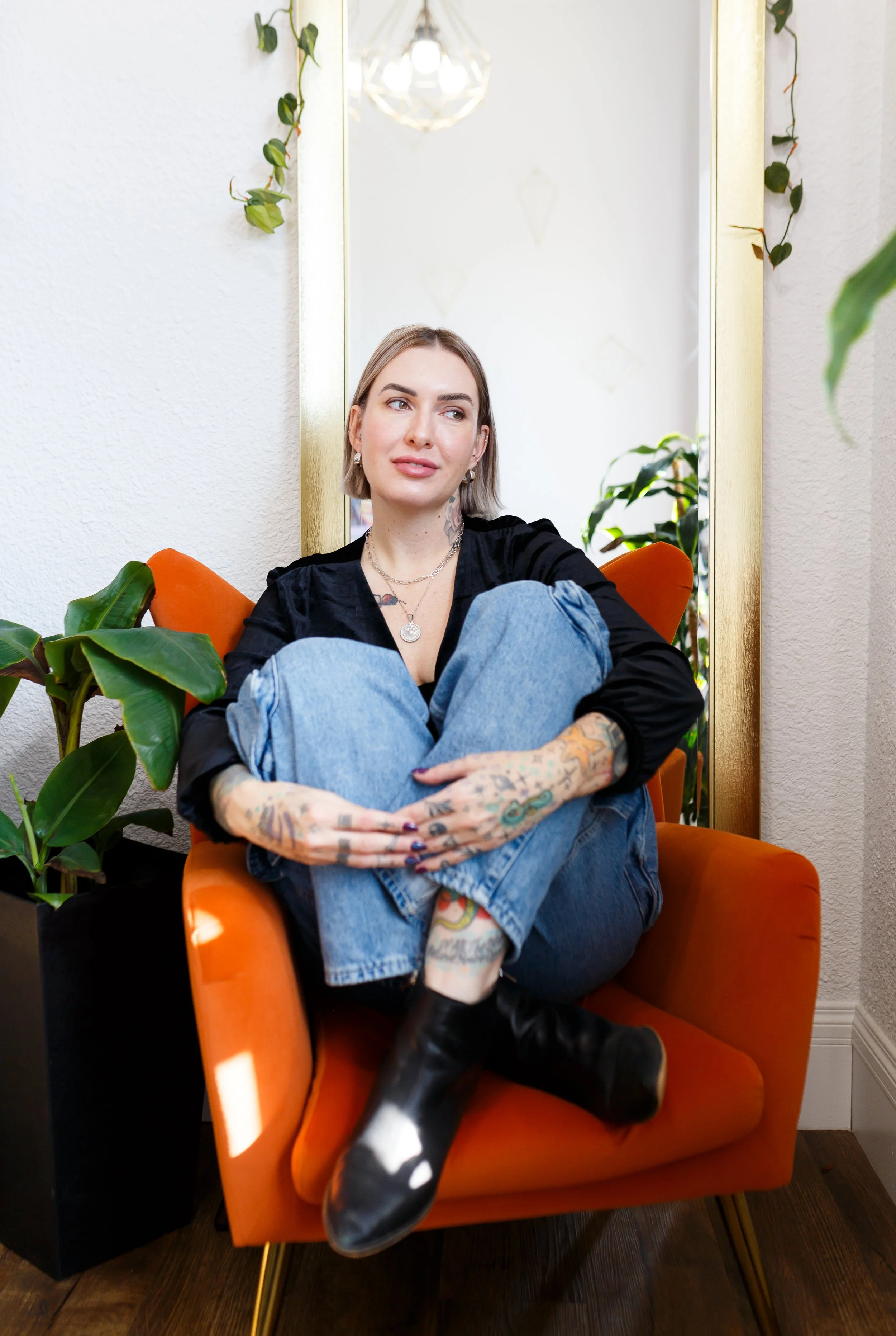Lashious Studio owner with tattoos sitting cross-legged in an orange armchair in front of a mirror, with plants around her, in an indoor space with white walls and wooden flooring.