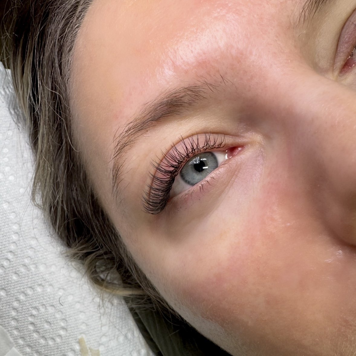 Close-up of a person's left eye with long eyelashes, light blue iris, and smooth skin, with a small portion of the nose and eyebrow visible.