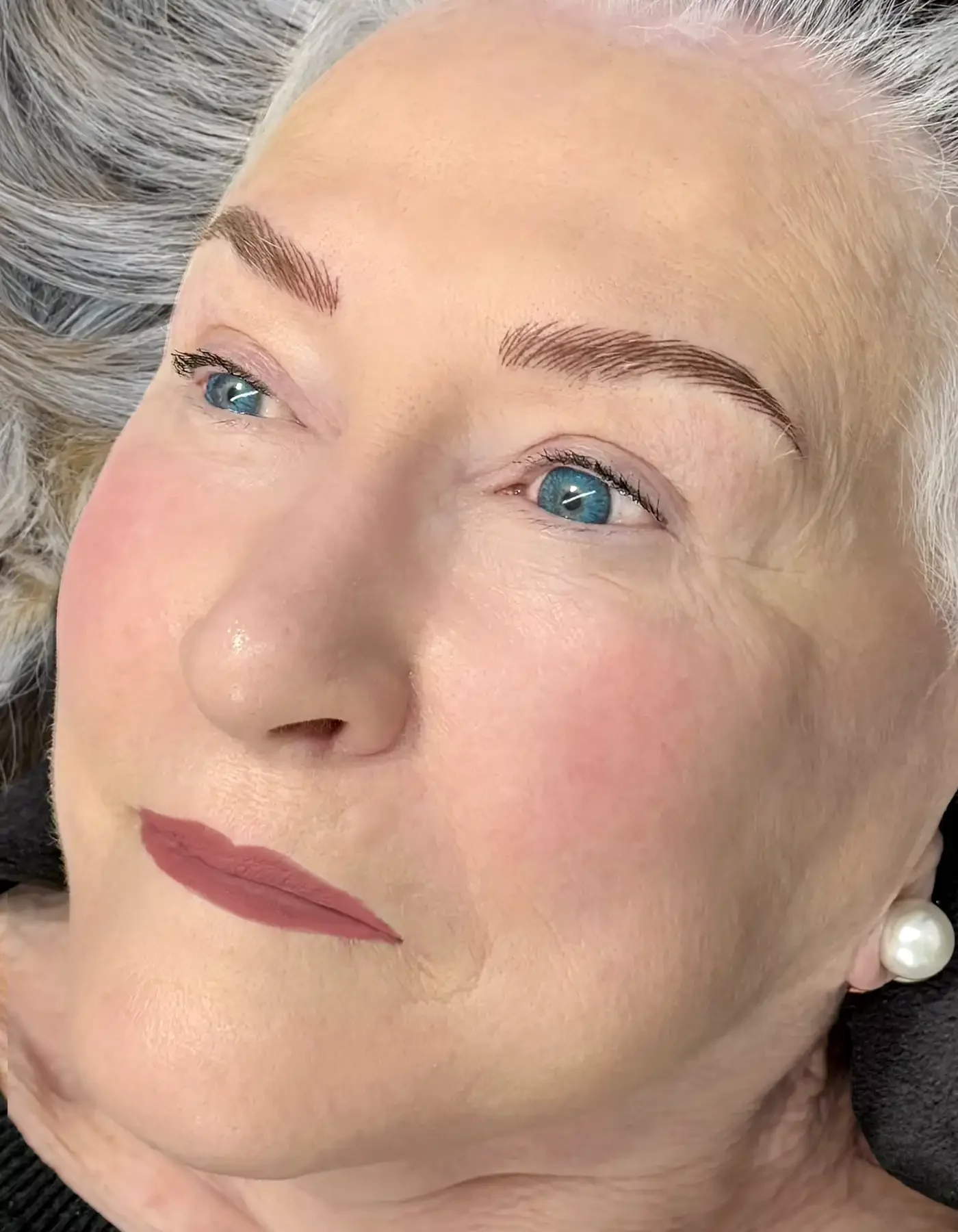 Close-up of a middle-aged woman with blue eyes and blonde hair. She has well-groomed eyebrows, soft makeup, and is wearing pearl earrings. Her face is turned slightly to the side, with a calm expression.
