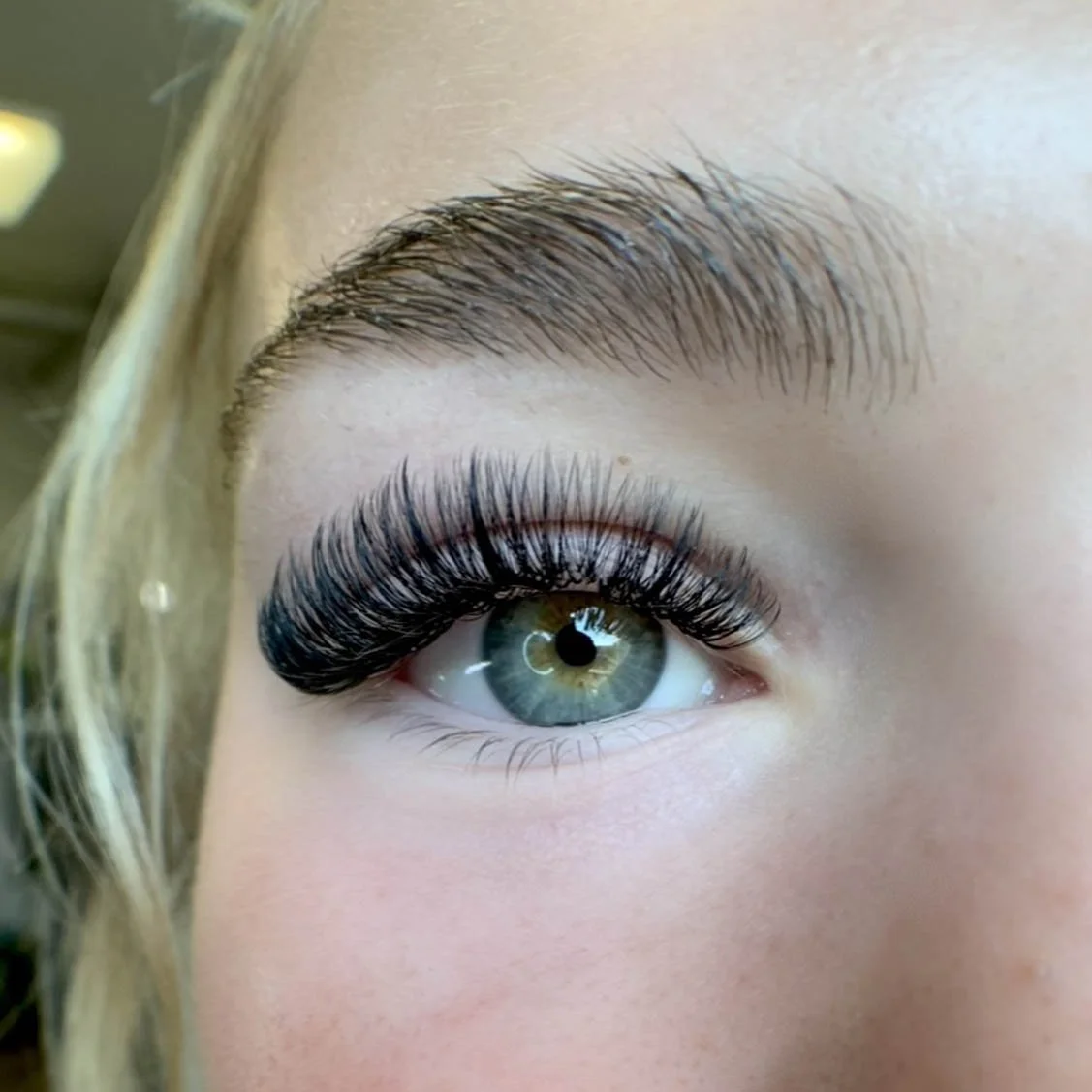 Close-up of a woman's eye with long, thick eyelashes and well-groomed eyebrow, green iris, and clear skin.
