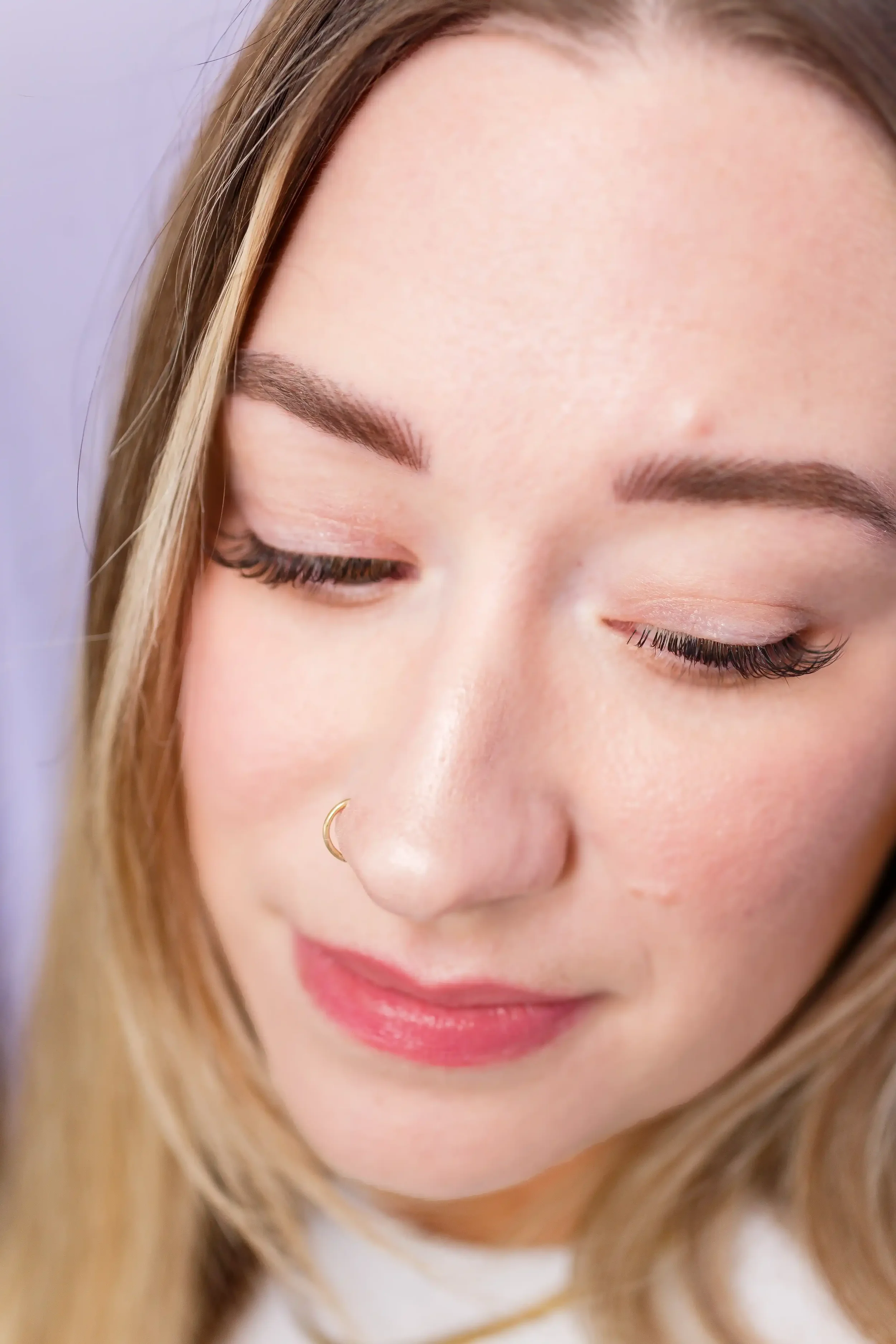 Close-up of a woman with light skin, closed eyes, long eyelashes, and a small nose ring, smiling softly.