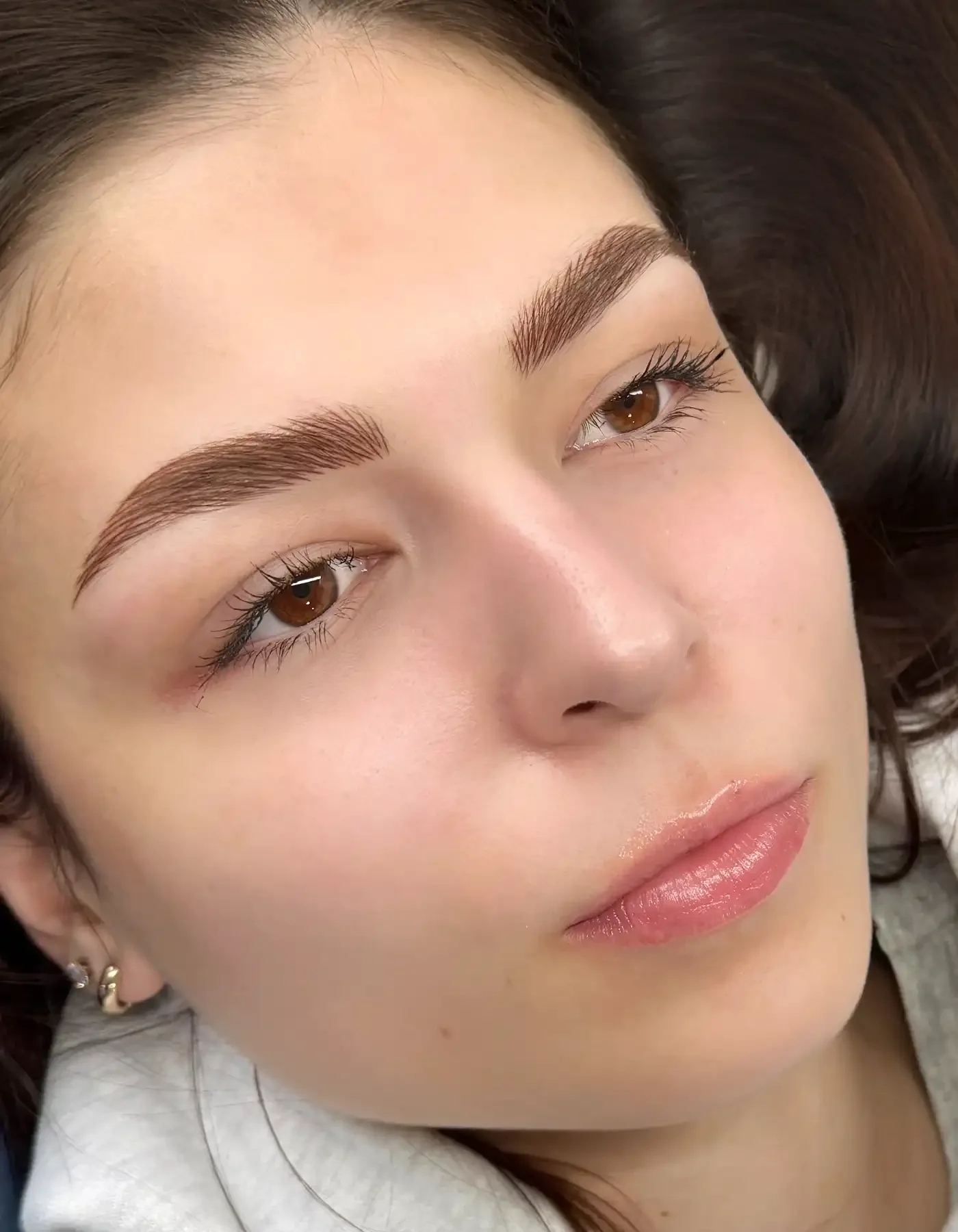 Close-up of a woman's face with well-groomed eyebrows, light makeup, and glossy lips, lying down with her head slightly tilted.