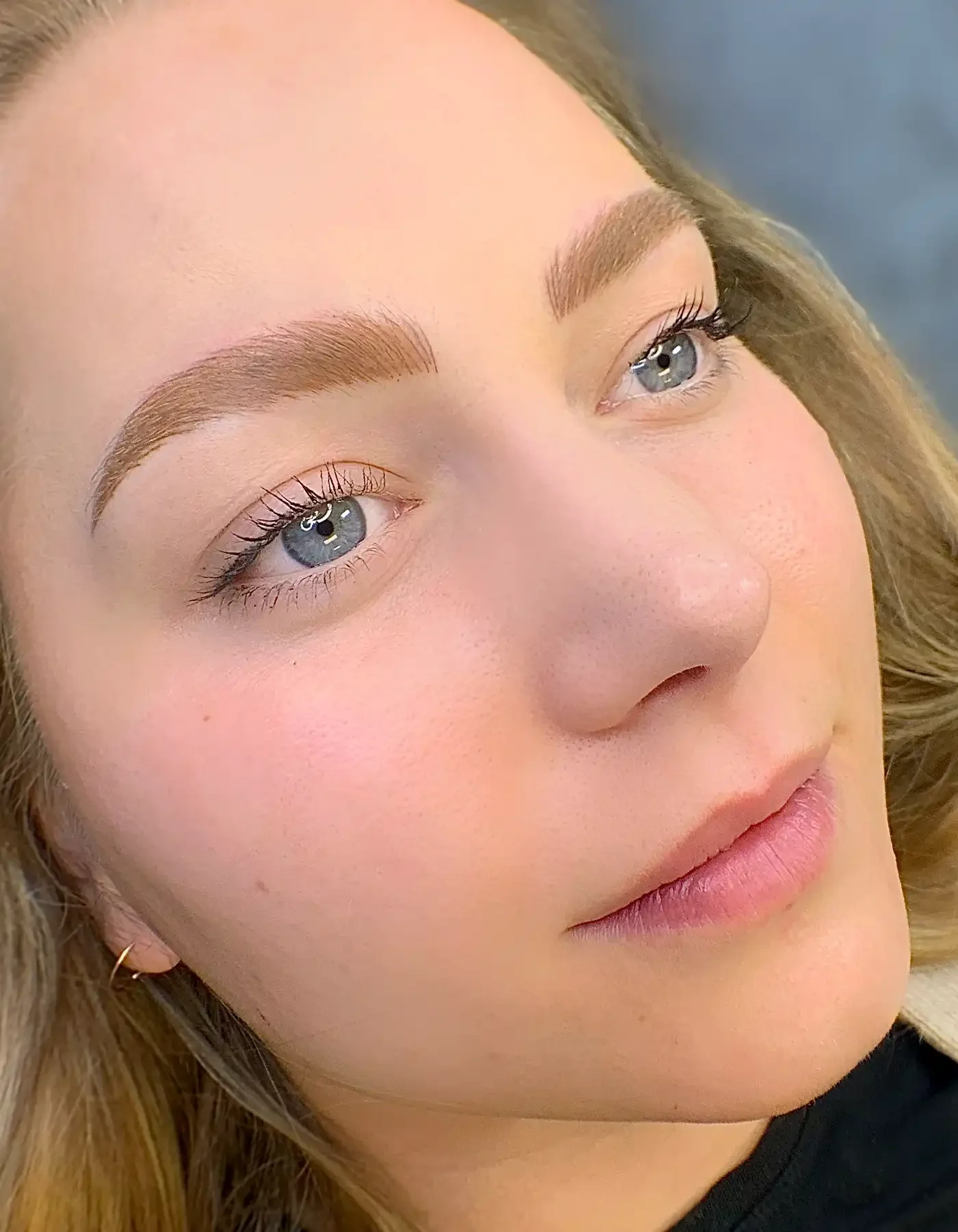 Close-up of a young woman's face with clear skin, light makeup, and nano brows. She has blue eyes and light brown hair.