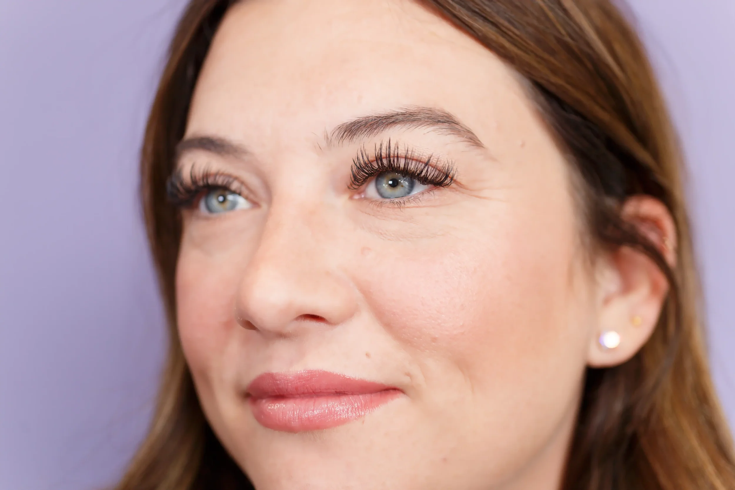 Close-up of a woman's face showing her blue eyes, permanent makeup, and earrings, with a light purple background.