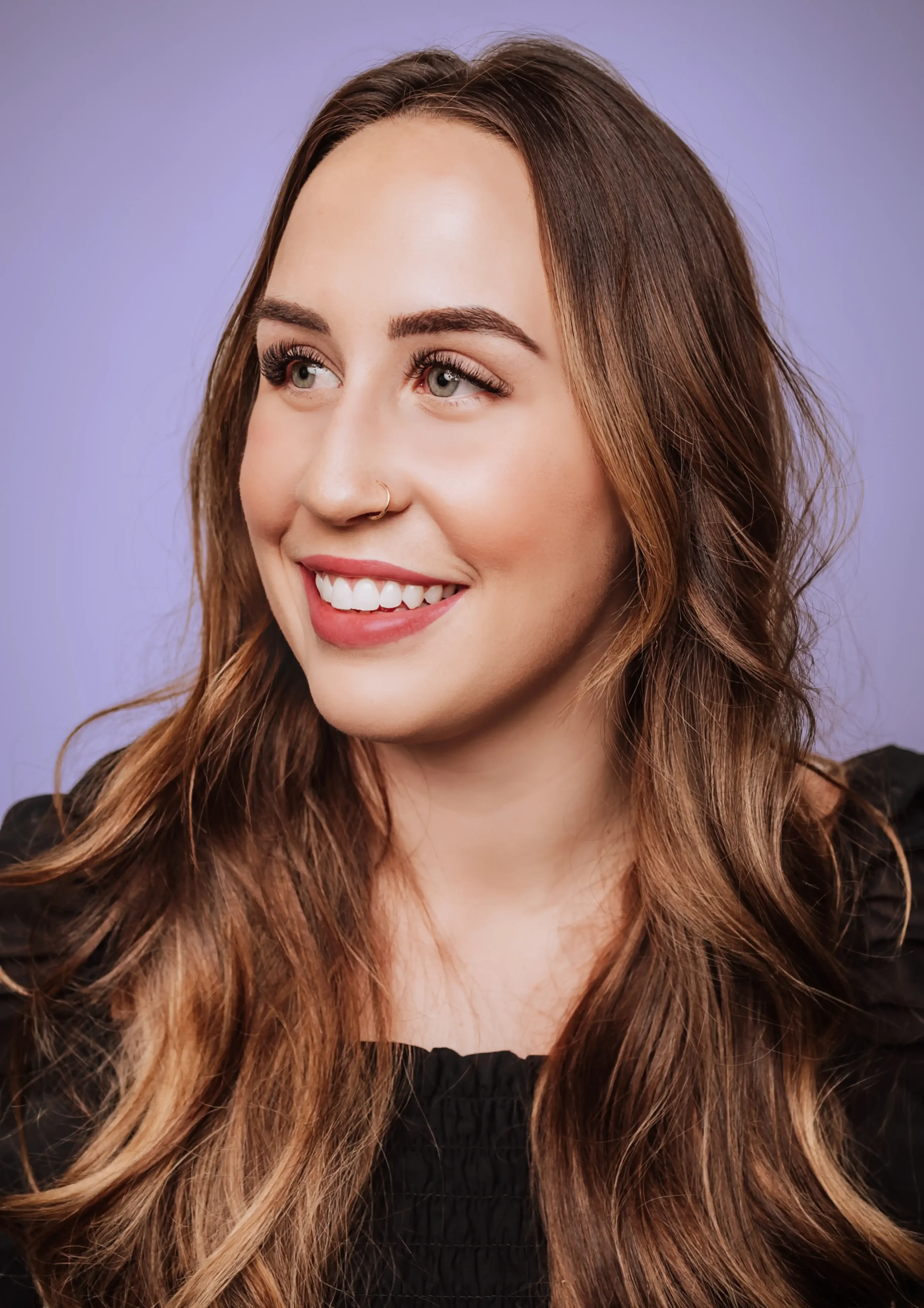 Portrait of a smiling young woman with brown wavy hair, light skin, wearing a black top, against a purple background.
