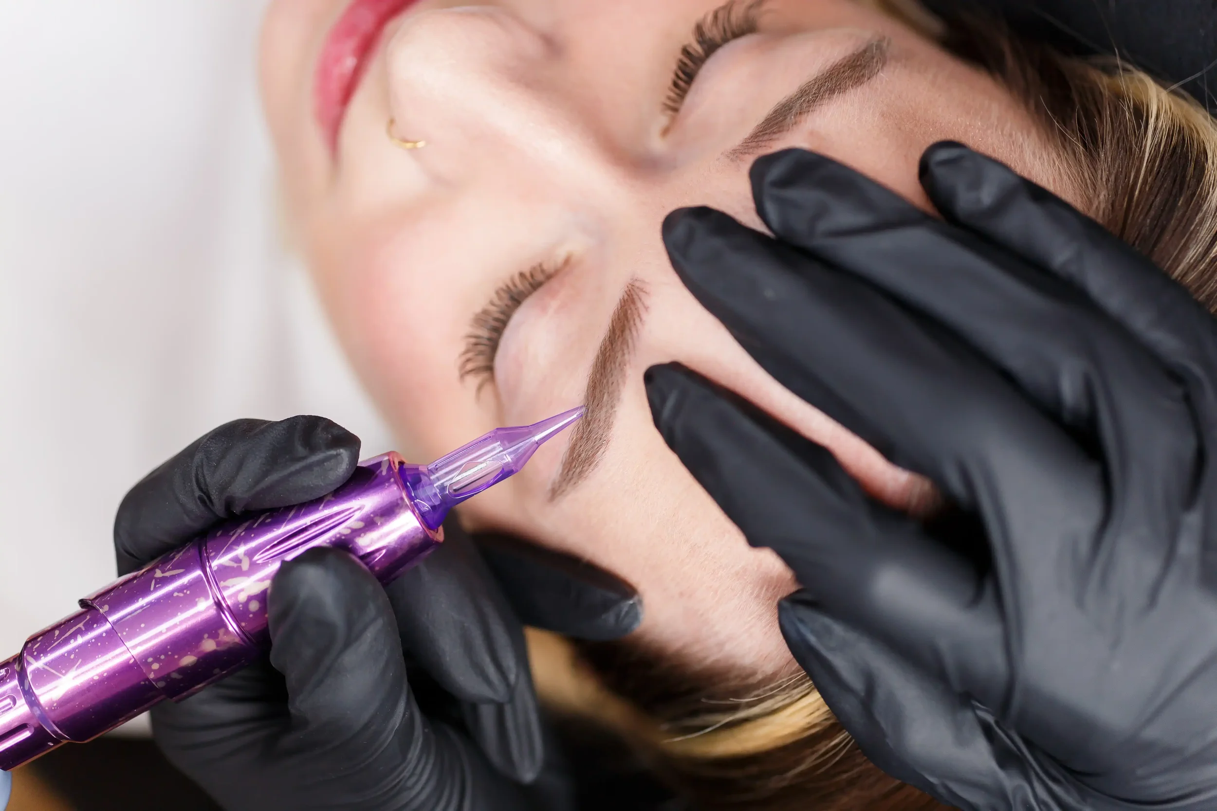 A person receiving eyebrow tattooing or microblading treatment, with a technician using a purple tattoo pen, while wearing black gloves.