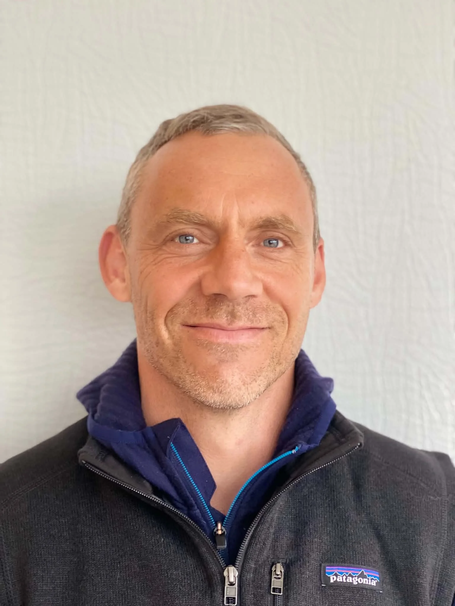 Close-up of a smiling man with blue eyes and short gray hair, wearing a Patagonia black fleece jacket with a purple and blue shirt visible underneath, standing against a plain white wall.