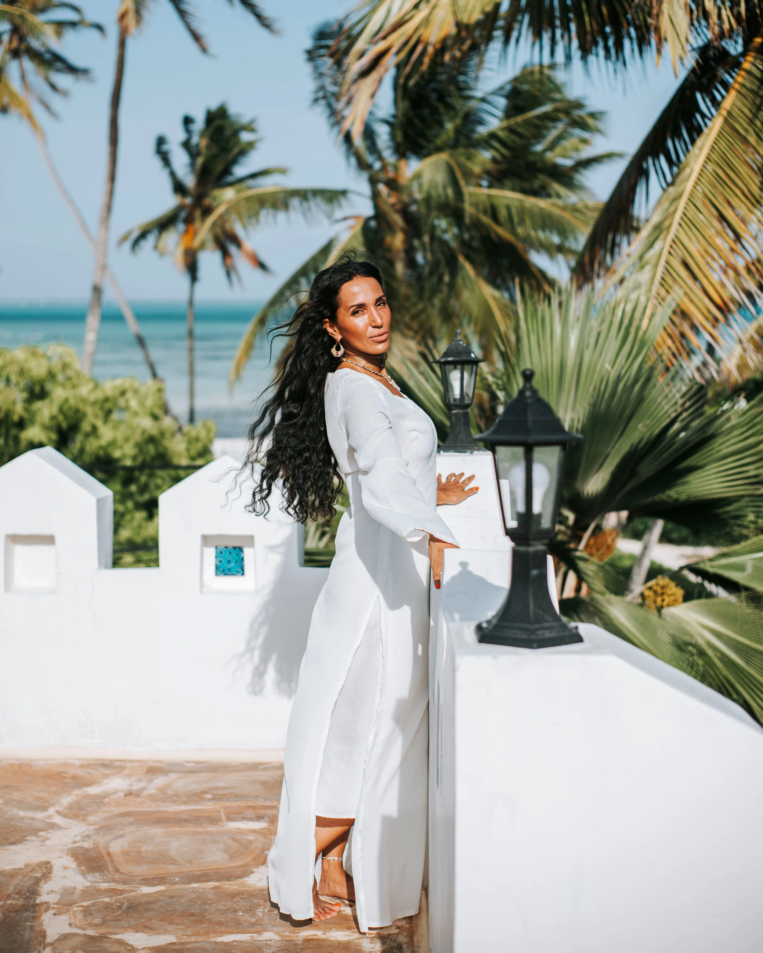 Woman in white dress standing near a white wall with black lanterns, overlooking a tropical beach with palm trees and ocean.