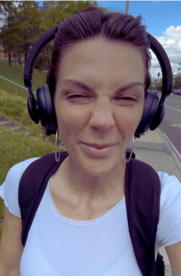 A woman with short brown hair wearing large black headphones, a white shirt, and a black backpack, smiling with her eyes closed outdoors on a sunny day near a street with trees and grass in the background.